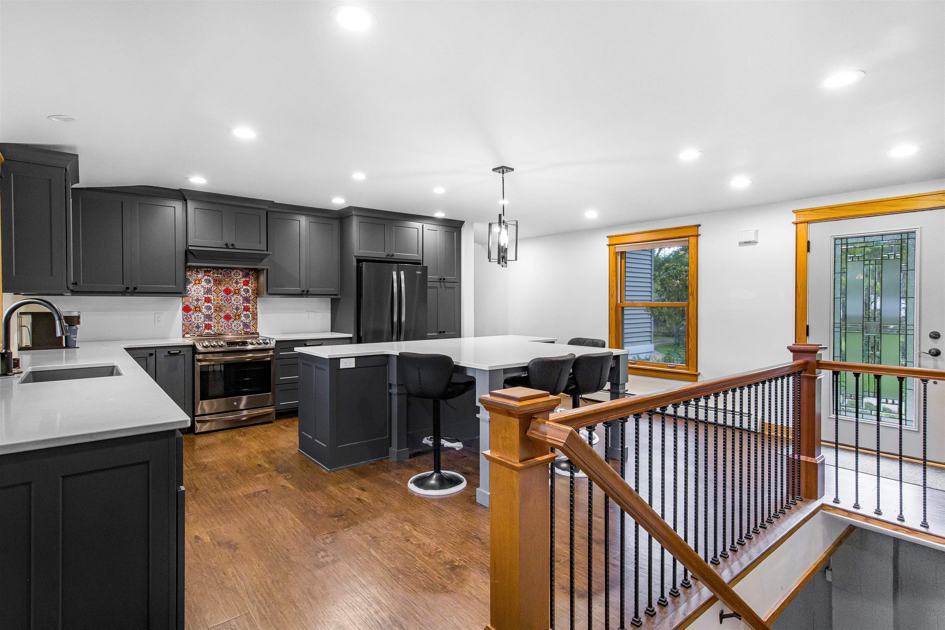 Open kitchen with dark cabinets, colorful backsplash, large island, and adjacent wood-railed staircase.