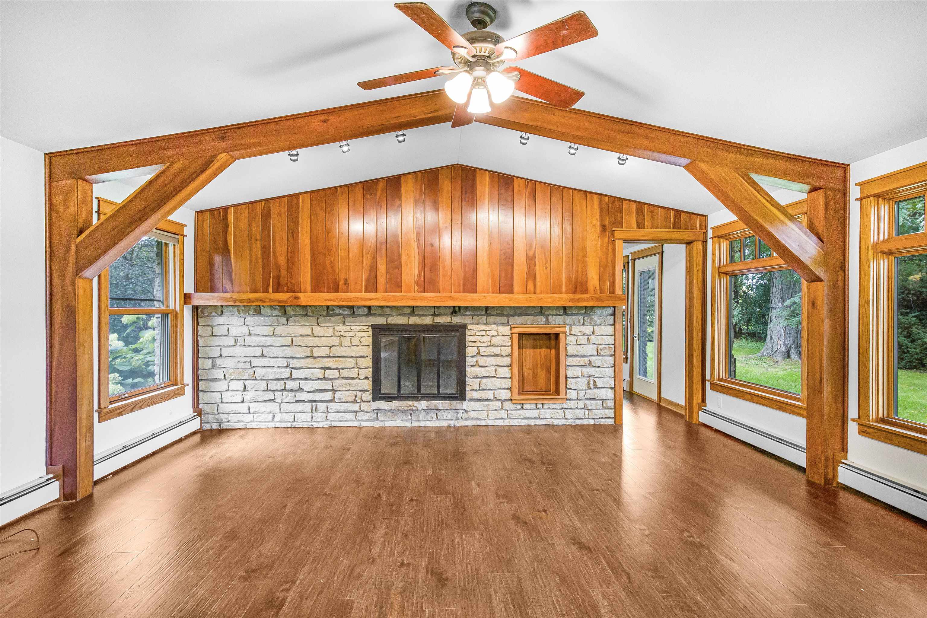 Empty vaulted living room with timber beams, a stone fireplace, wood paneling, and large windows.