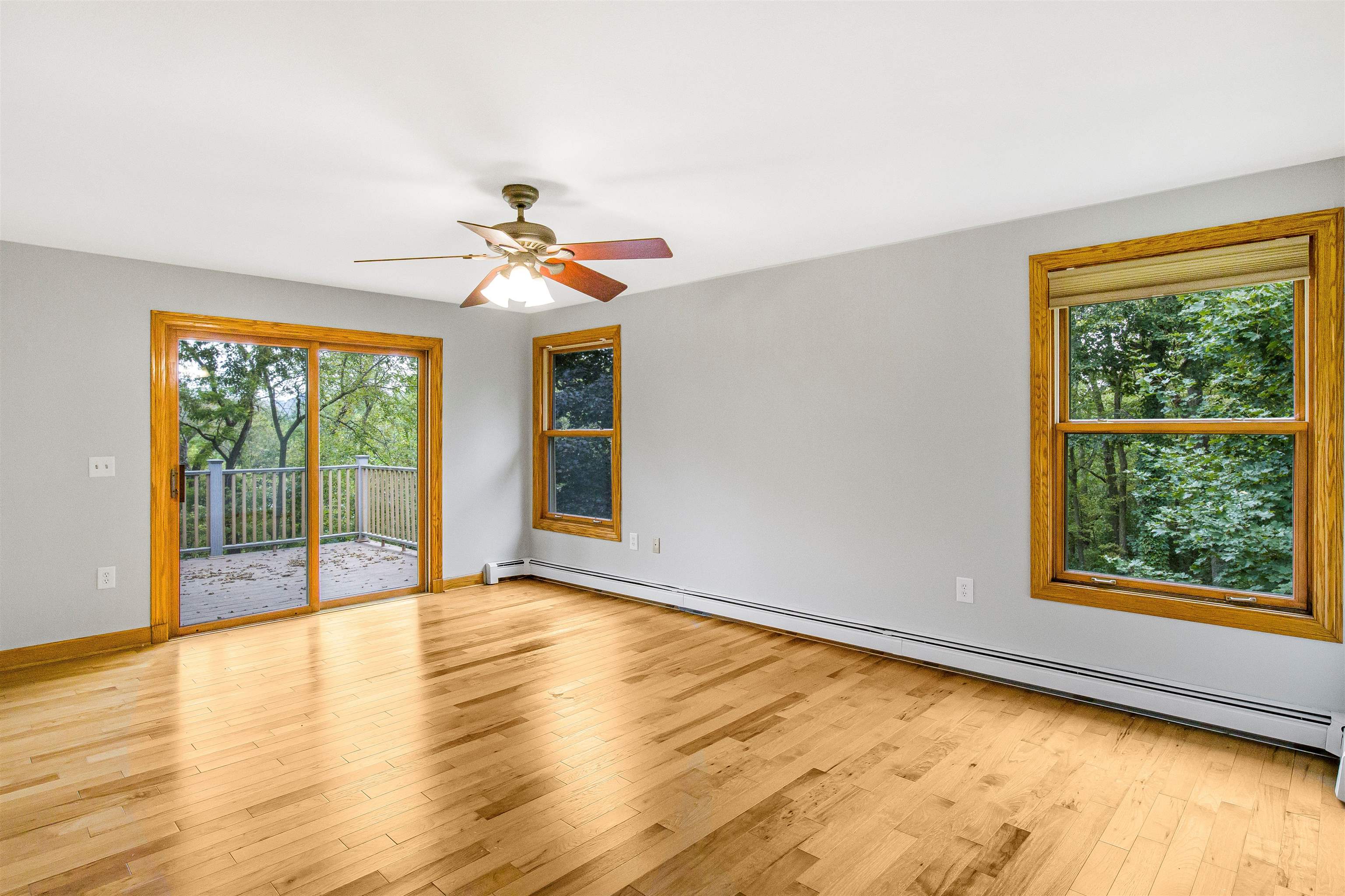 Empty room with hardwood floors, a sliding glass door to a deck, and wooded views through wood-trimmed windows.