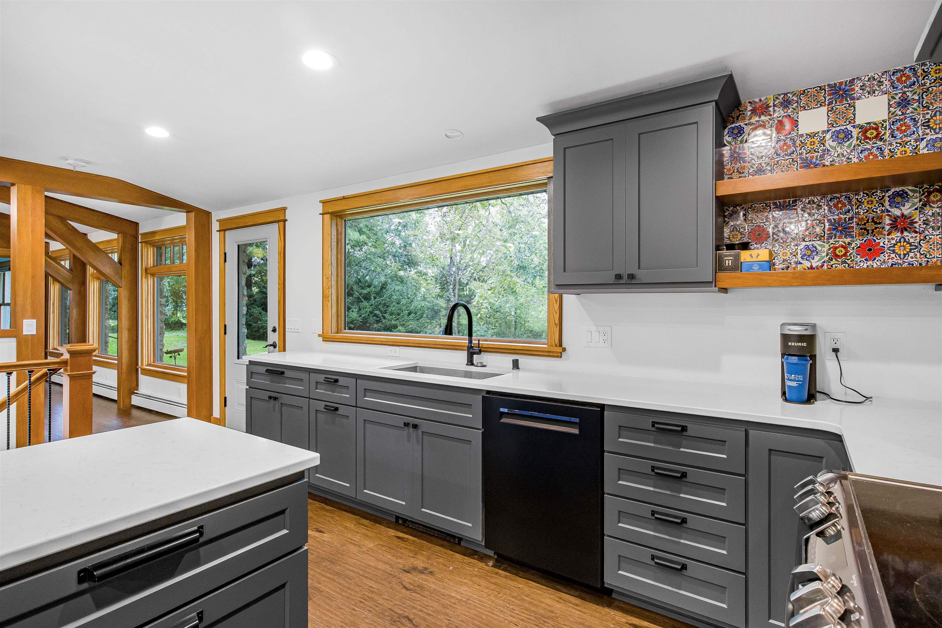 Kitchen with gray cabinets, colorful patterned tile backsplash, and a large window overlooking trees.