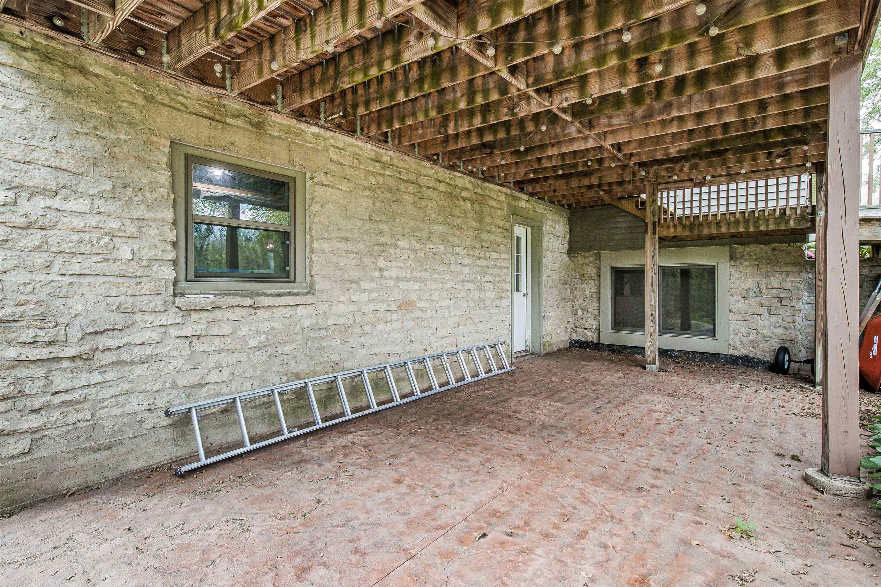 Covered stone patio beneath a raised wooden deck, with string lights and a ladder against the wall.