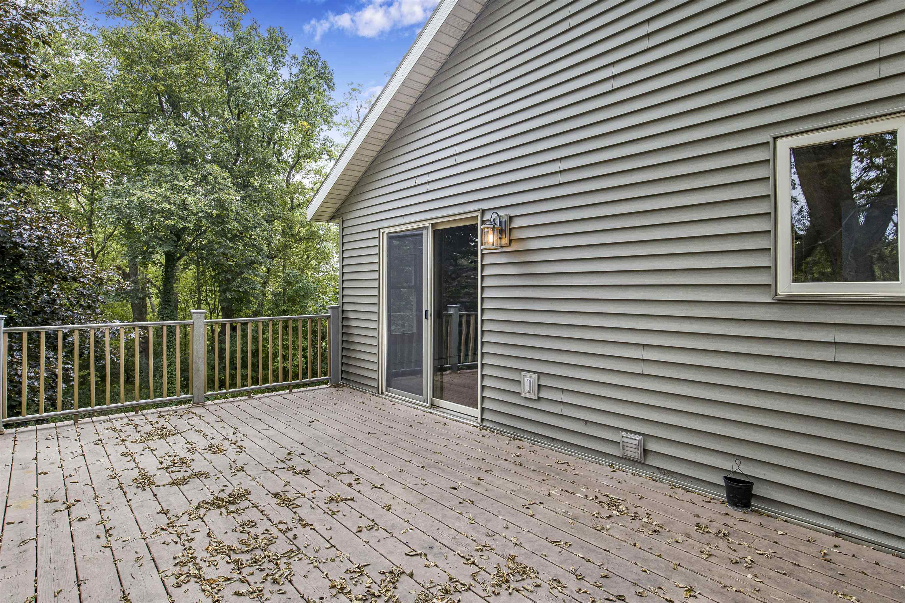 Wooden deck beside a gray-sided home, with a sliding door and leafy trees beyond the railing.
