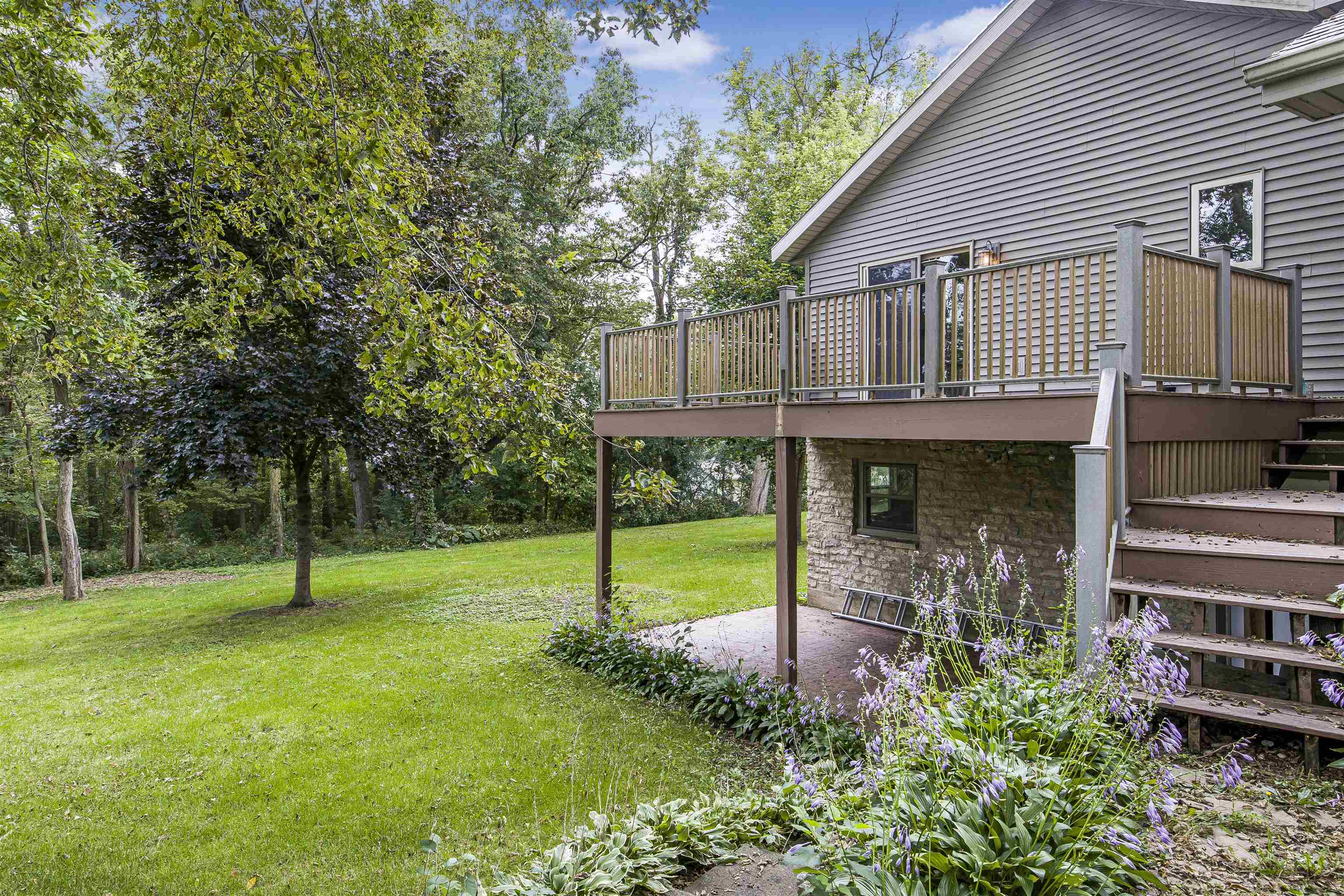 Home’s elevated deck and wooden stairs beside a green lawn, mature trees, and purple flowers.