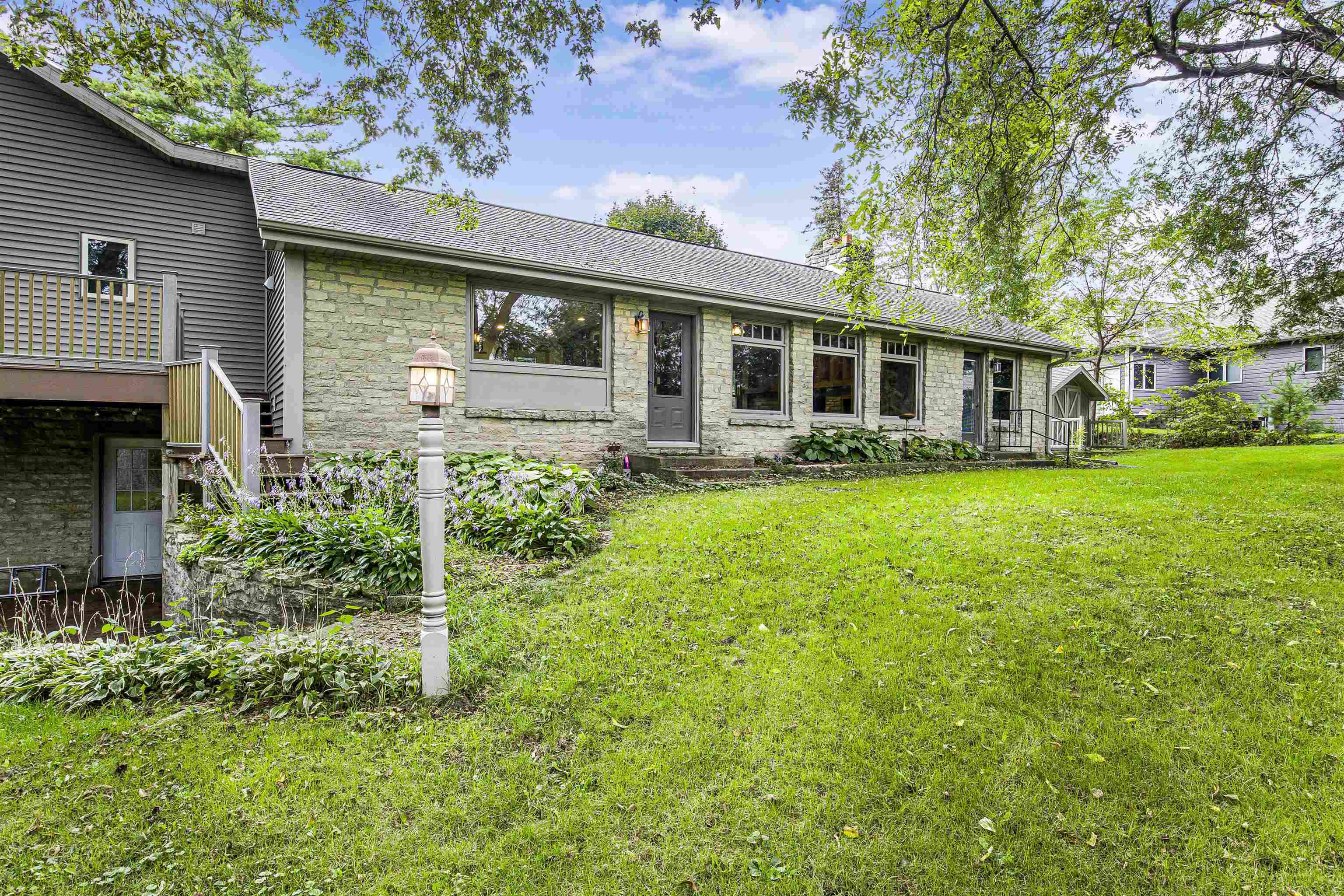 Long stone-and-gray-sided home with a raised deck, garden beds, and a broad green lawn.