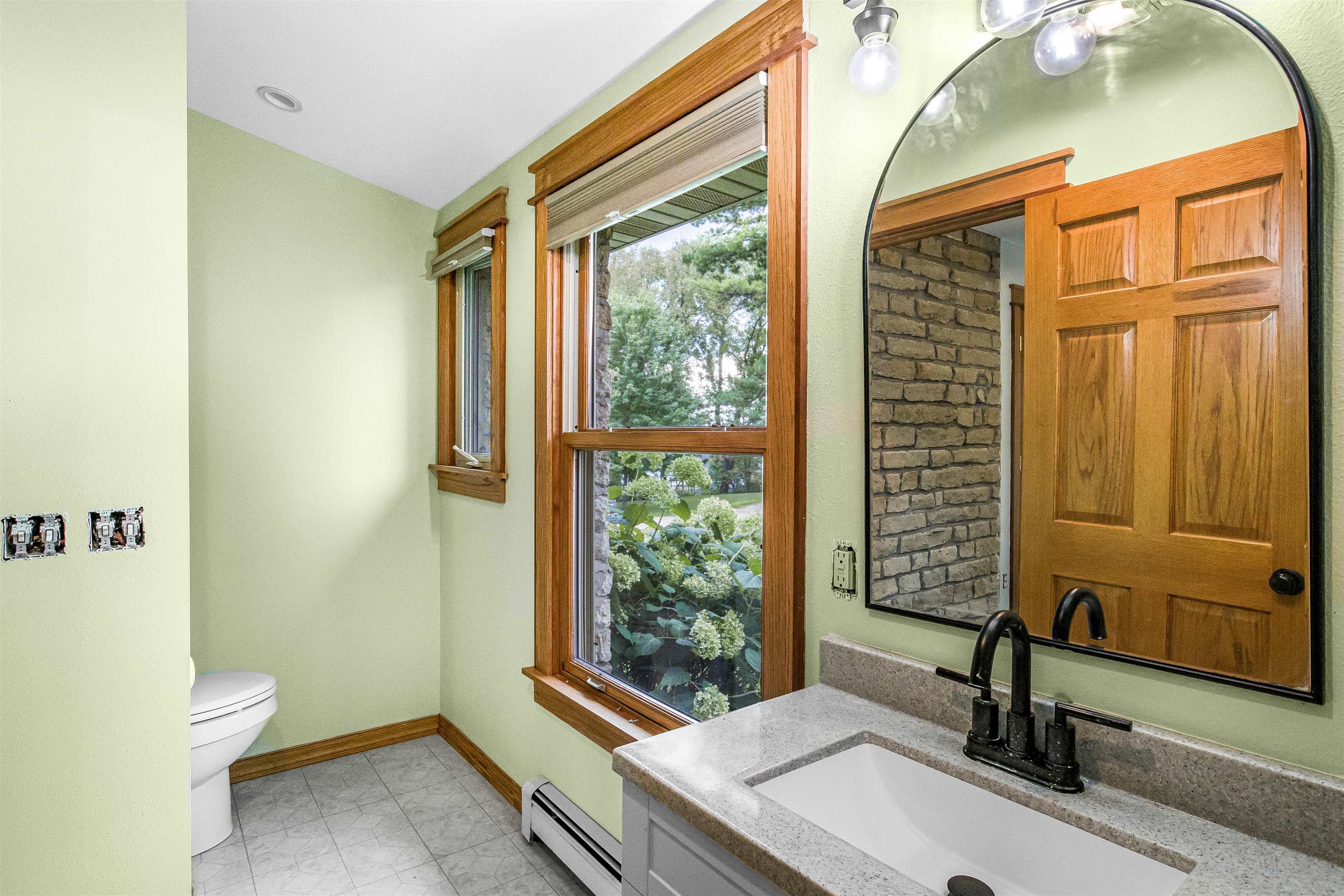 Bathroom with wood-trimmed garden-view windows, a stone-countertop vanity, and an arched mirror.