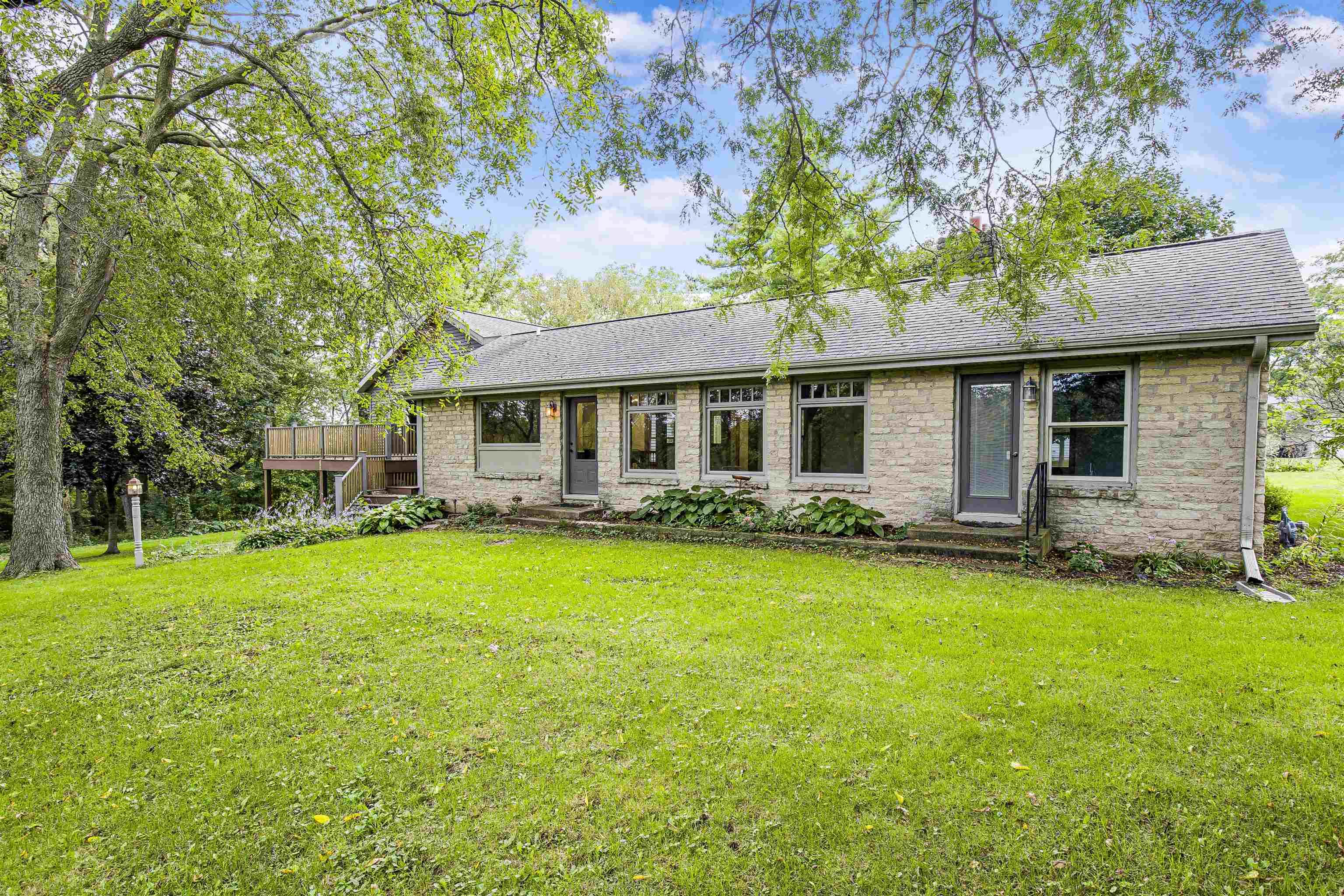 Single-story stone house with a wooden deck and broad lawn beneath mature trees.