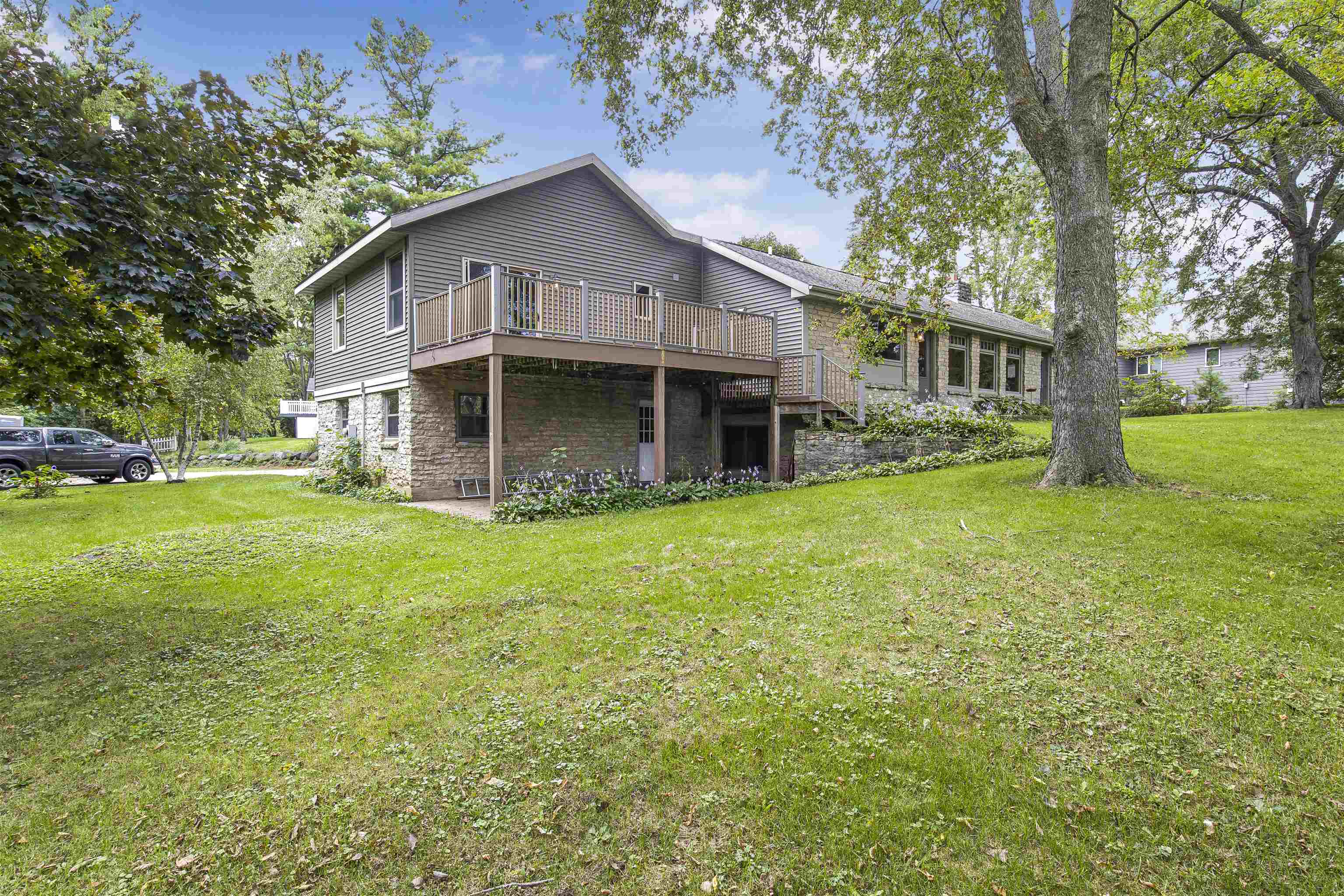 Rear exterior of a gray split-level house with a raised deck and stone lower level, surrounded by a leafy lawn.