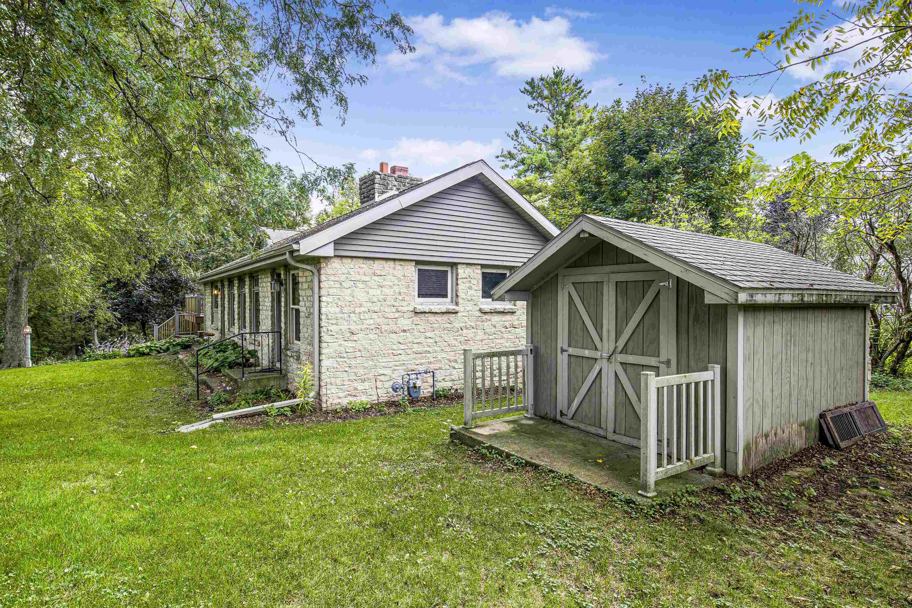 Stone house and gray gabled shed amid mature trees and a green lawn.