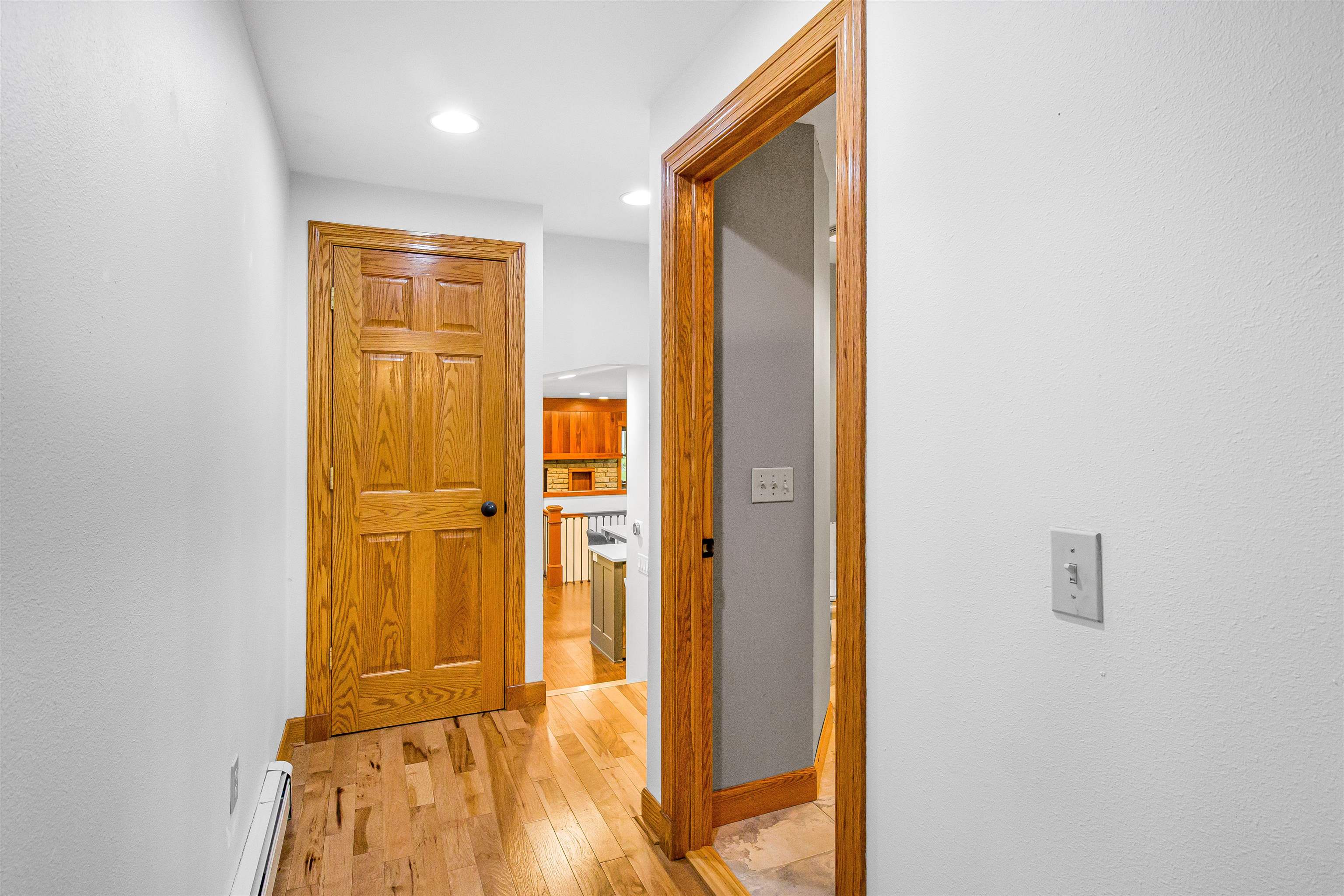 Hallway with hardwood floors and wood trim, opening toward a kitchen and stone-surround fireplace.