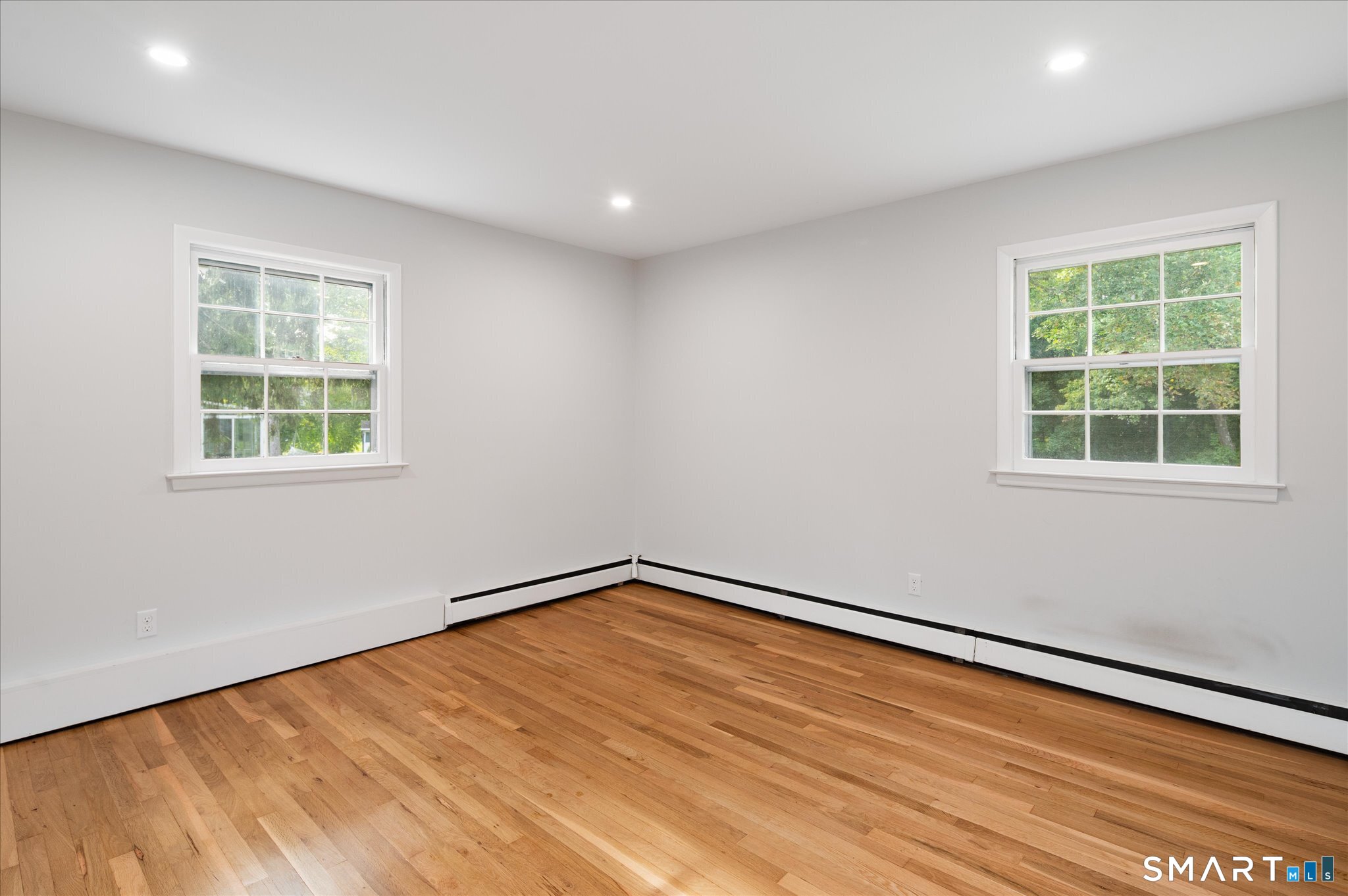Empty room with hardwood floors, two windows, recessed lights, and baseboard heating.