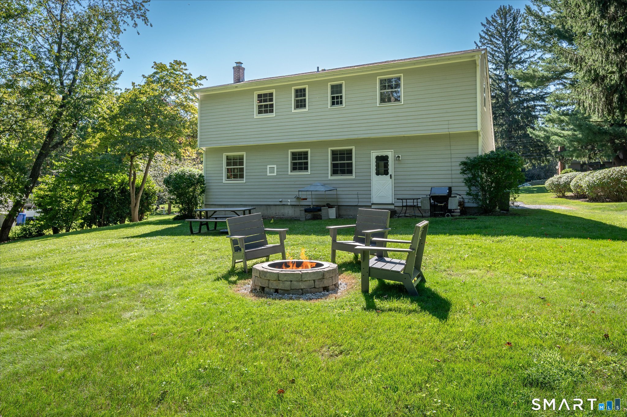Backyard with a stone fire pit, Adirondack chairs, broad lawn, and two-story gray-sided house.