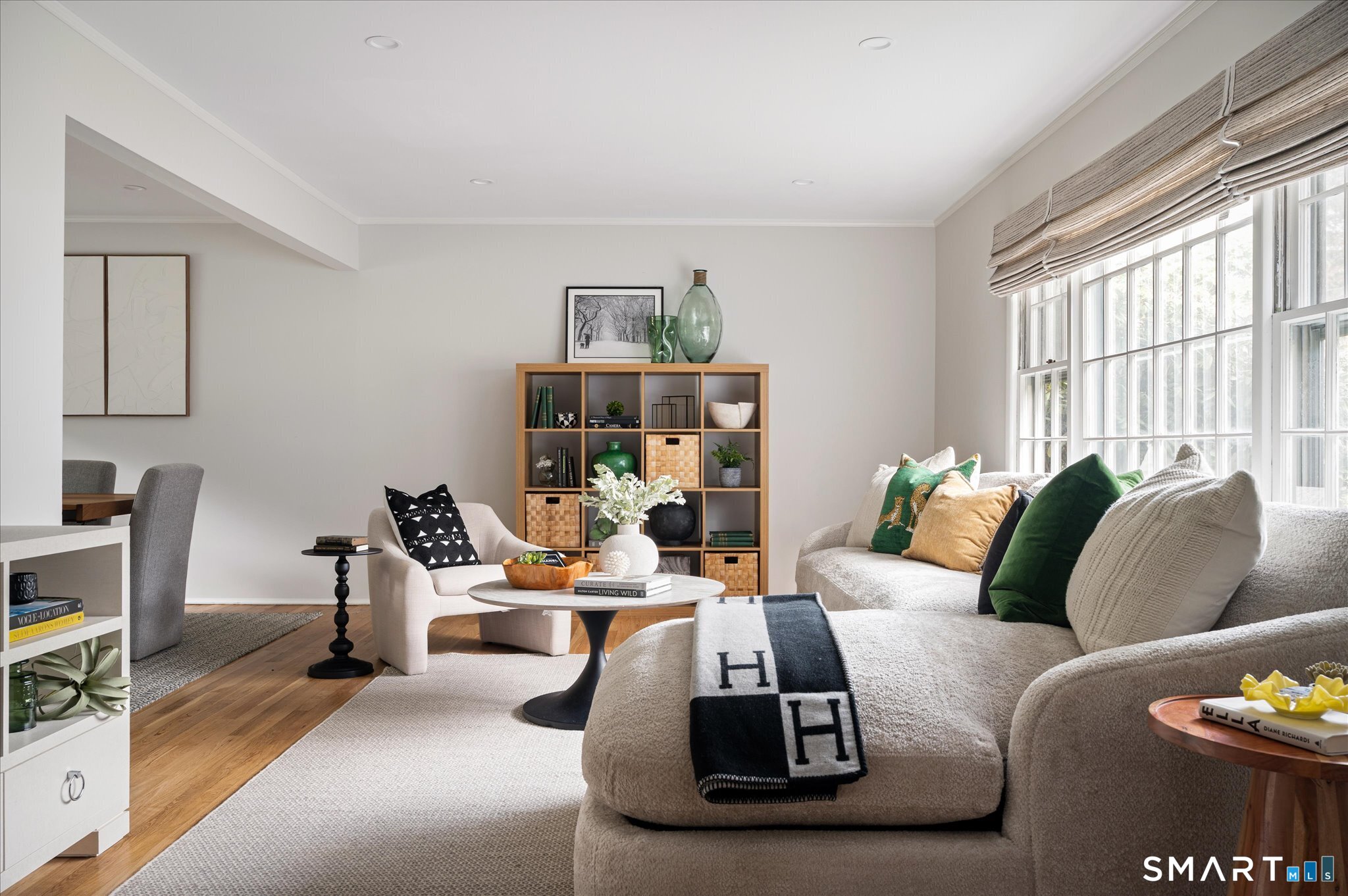Staged living room with a large sectional, green pillows, wood cubby bookcase, and tall bright windows.