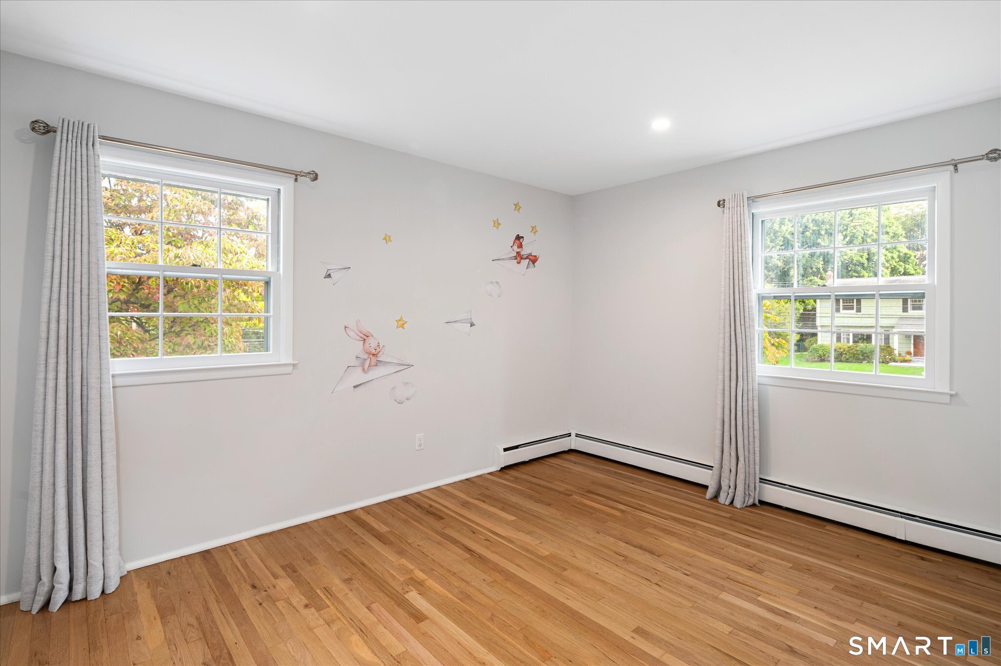 Empty bedroom with hardwood floors, two curtained windows, and whimsical wall decals.