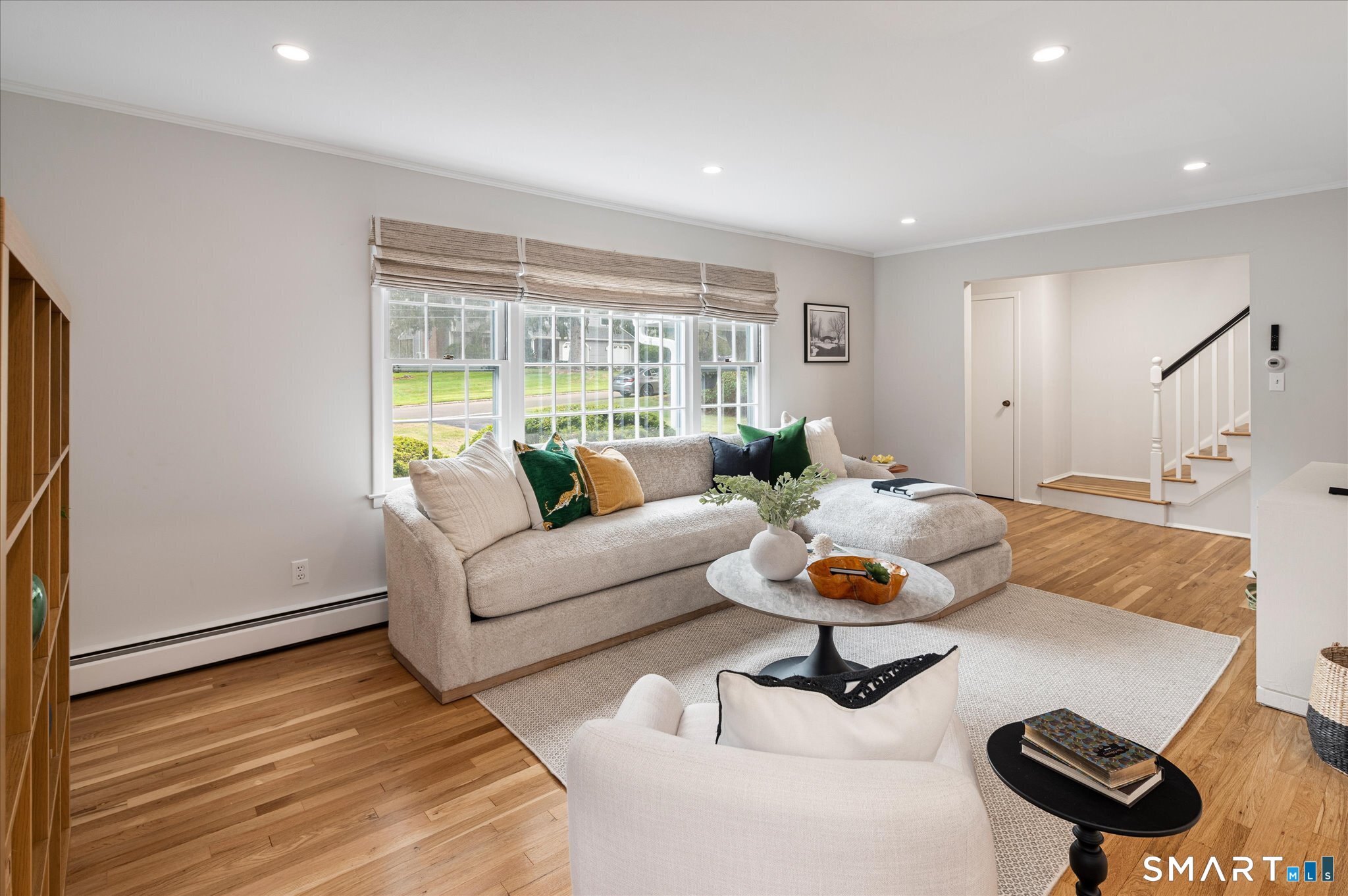 Staged living room with a beige sectional, hardwood floors, and large street-facing windows.