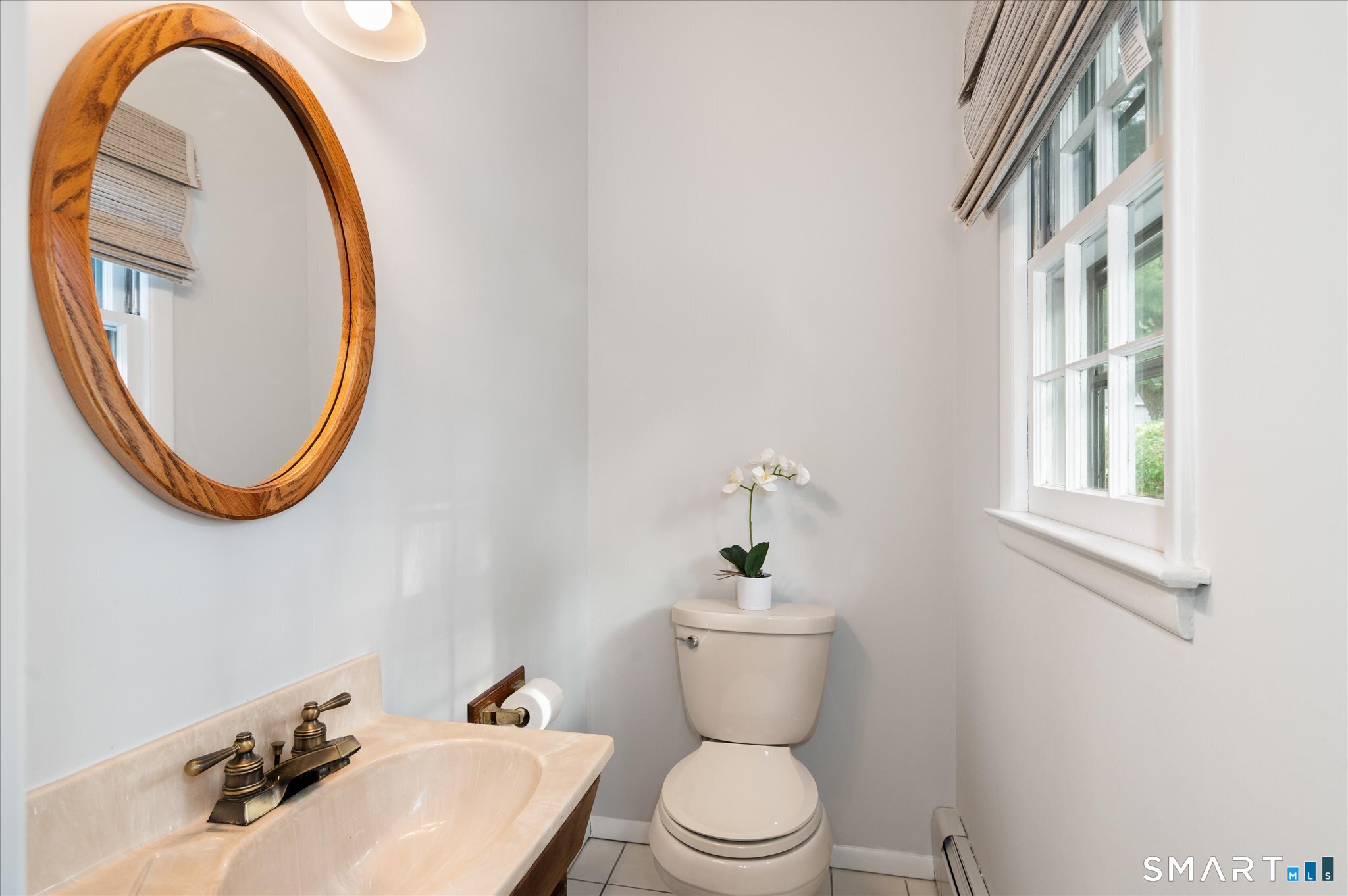 Half bathroom with a cream vanity, oval wood-framed mirror, and window with woven shades.