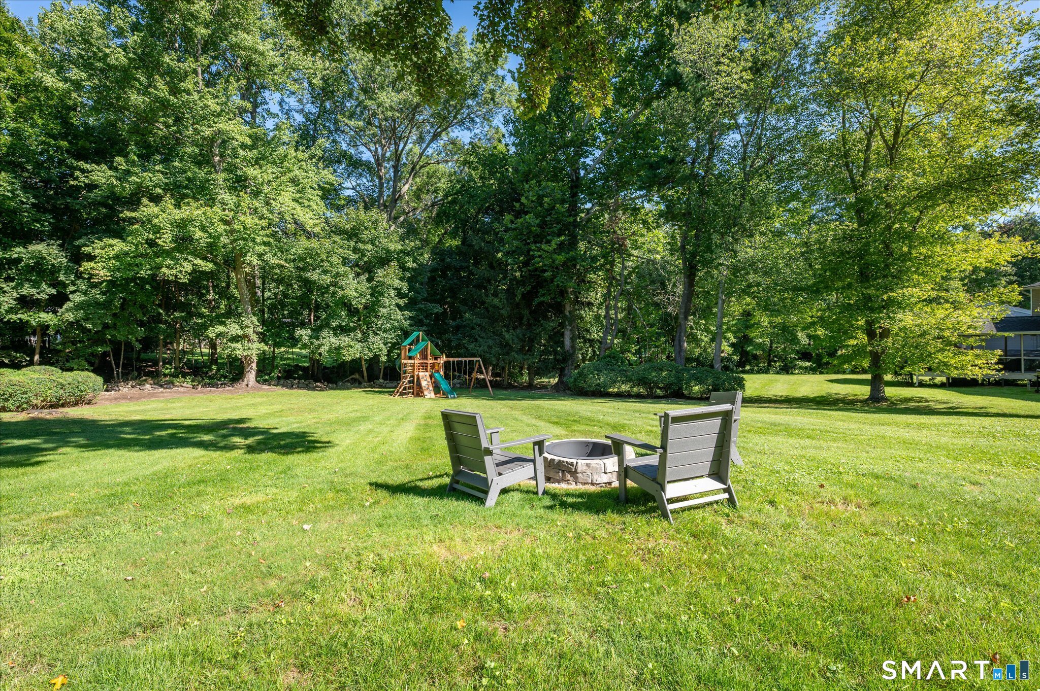 Spacious tree-lined backyard with a stone fire pit, two chairs, and a children’s playset.