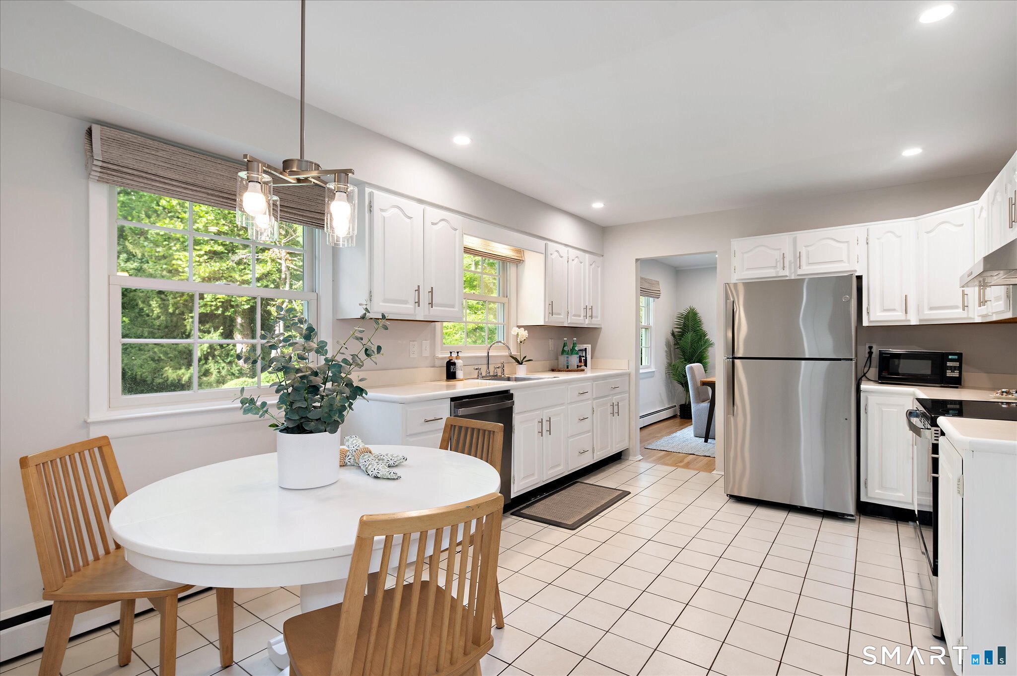 Bright white kitchen with stainless steel appliances, tiled floors, and a round dining table by large windows.