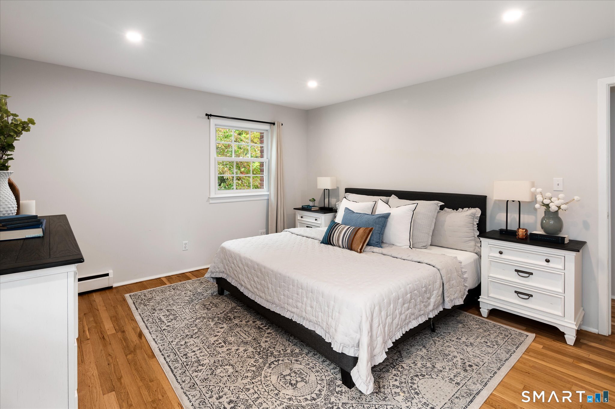 Staged bedroom with a white bed, black headboard, hardwood floors, and a large patterned rug.