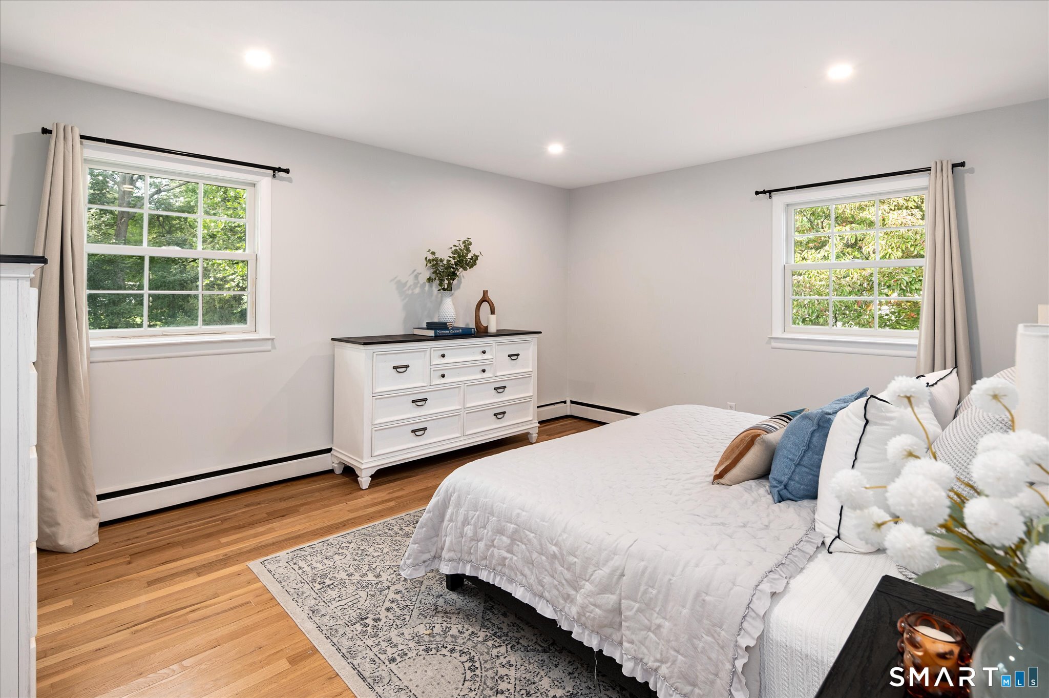 Staged bedroom with a white bed, hardwood floors, and two windows overlooking greenery.