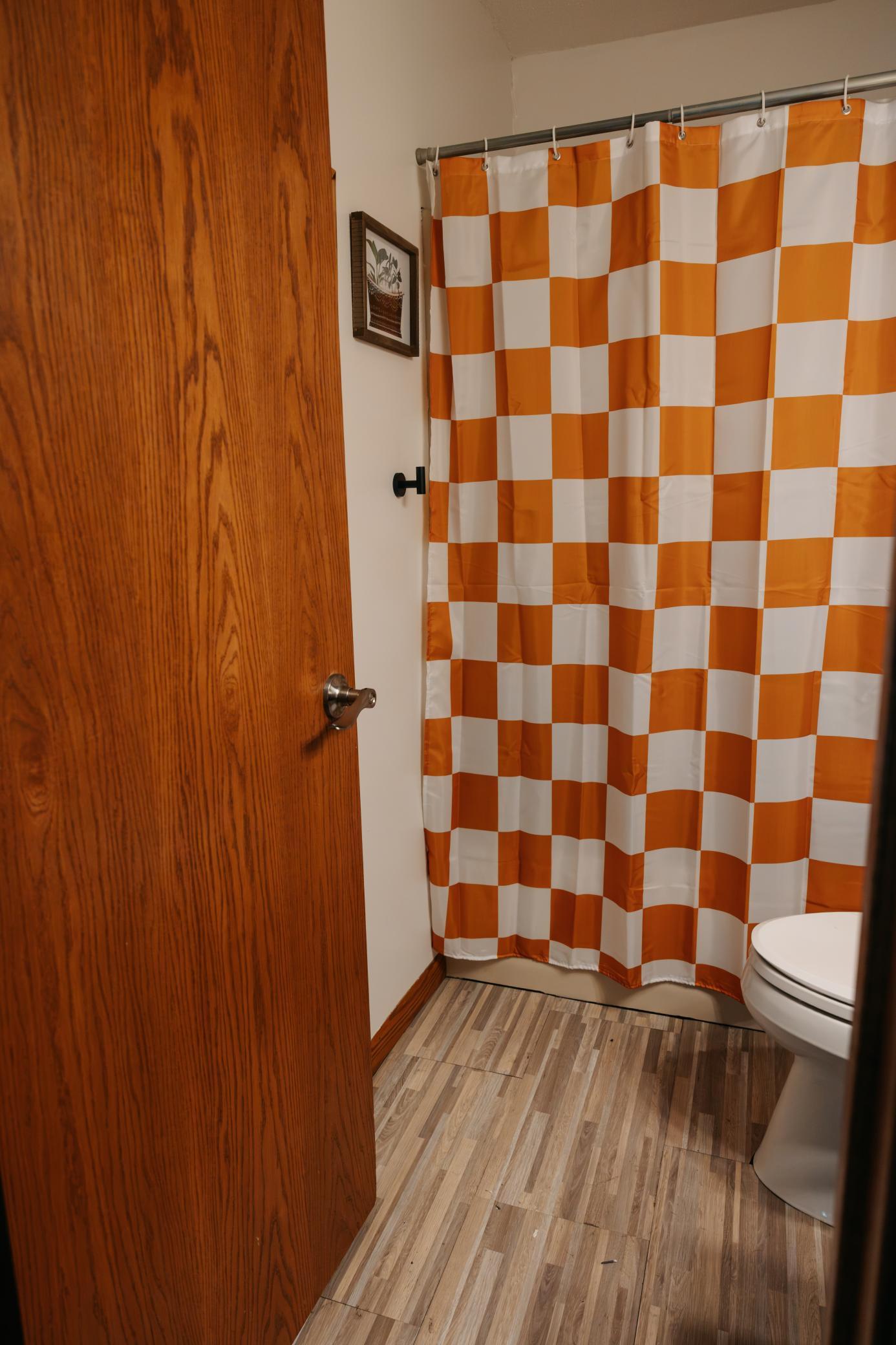 Bathroom with an orange-and-white checkerboard shower curtain and wood-look tile flooring.