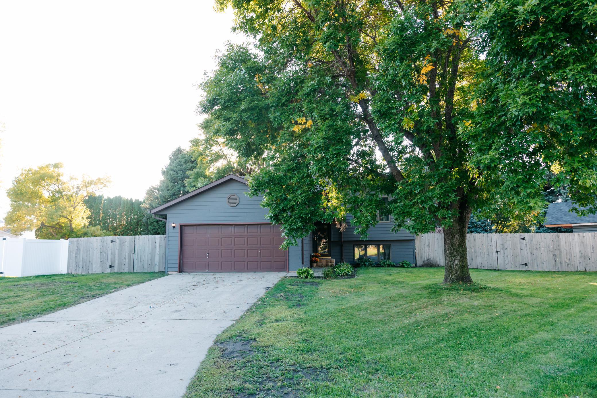Blue-gray split-level home with a double garage, mature shade tree, fenced lawn, and concrete driveway.
