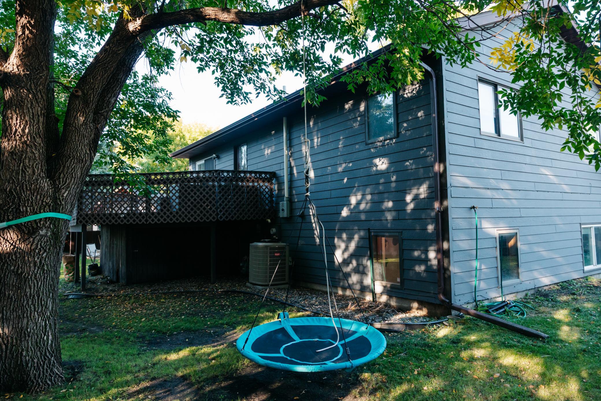 Rear of a blue-gray house with a raised deck, large shade tree, and turquoise circular swing.