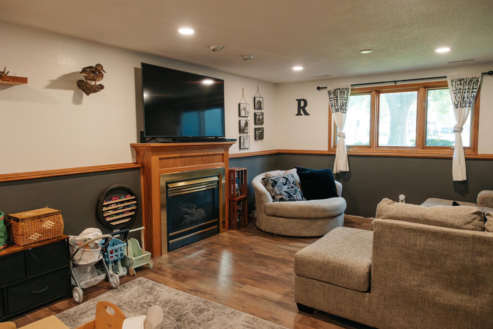 Living room with a wood-trimmed fireplace, large TV, beige seating, and wide curtained window.