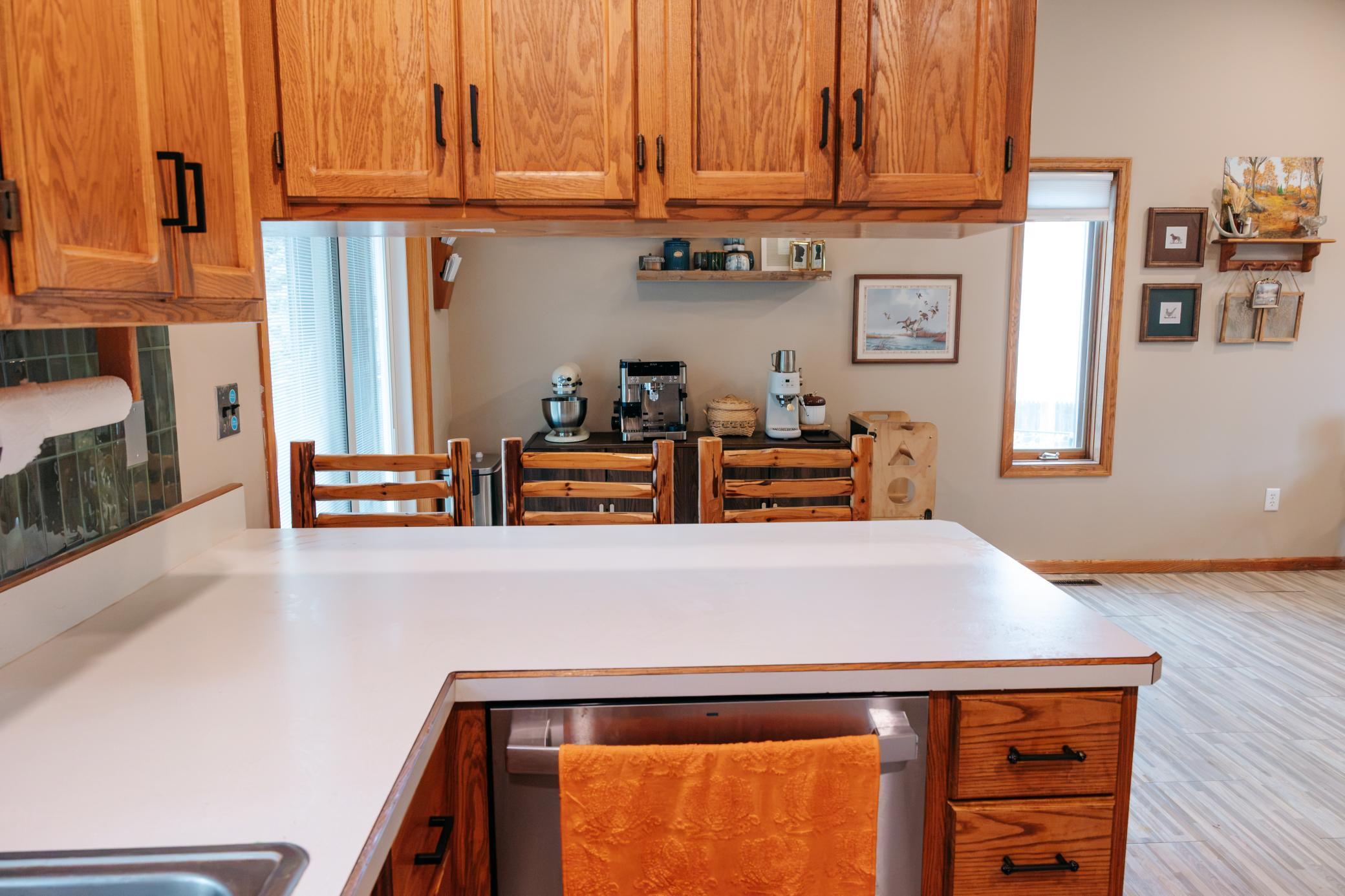 Kitchen with warm wood cabinets, white peninsula countertop, integrated dishwasher, and coffee station.
