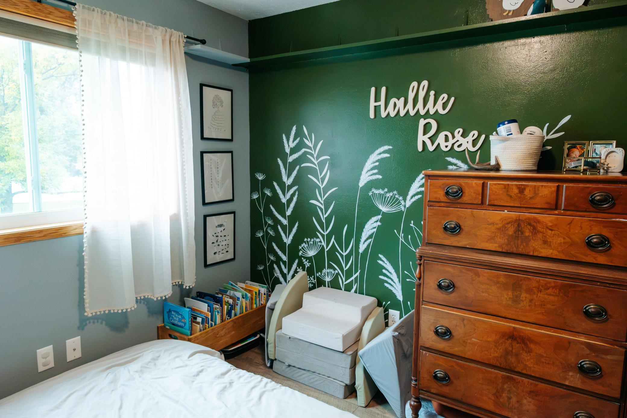Child’s bedroom with a green botanical accent wall, “Hallie Rose” lettering, and wooden dresser.