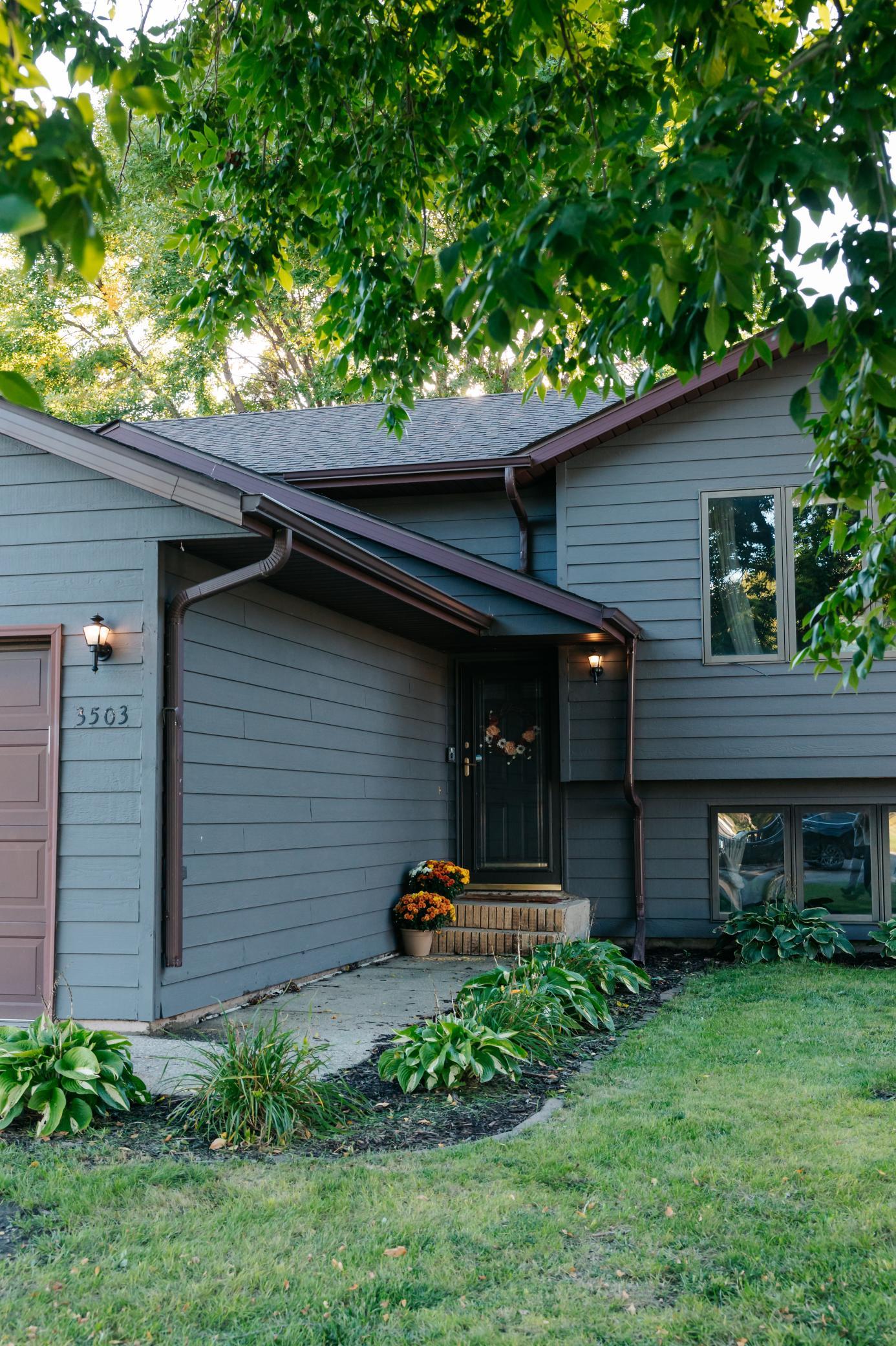 Gray-blue home entrance with dark trim, flowering planters, and leafy trees overhead.