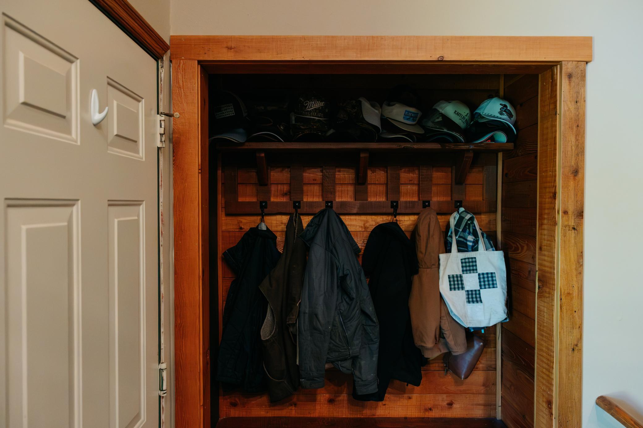 Wooden entryway closet with coats, caps, and a plaid tote bag.