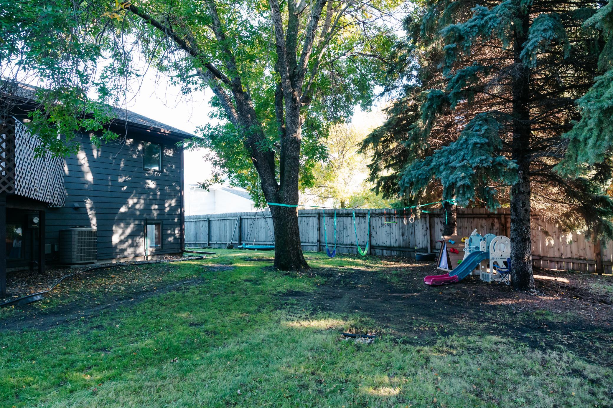 Fenced backyard with mature trees, swings, and a children's playset beside the house.
