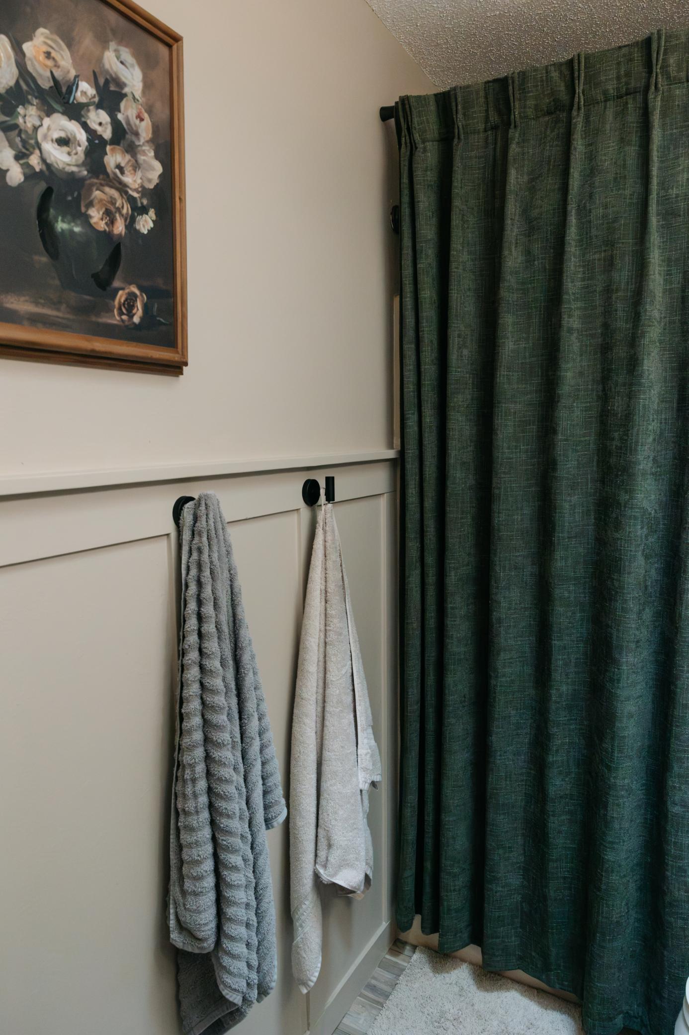 Bathroom corner with dark green shower curtains, hanging towels, and framed floral artwork.