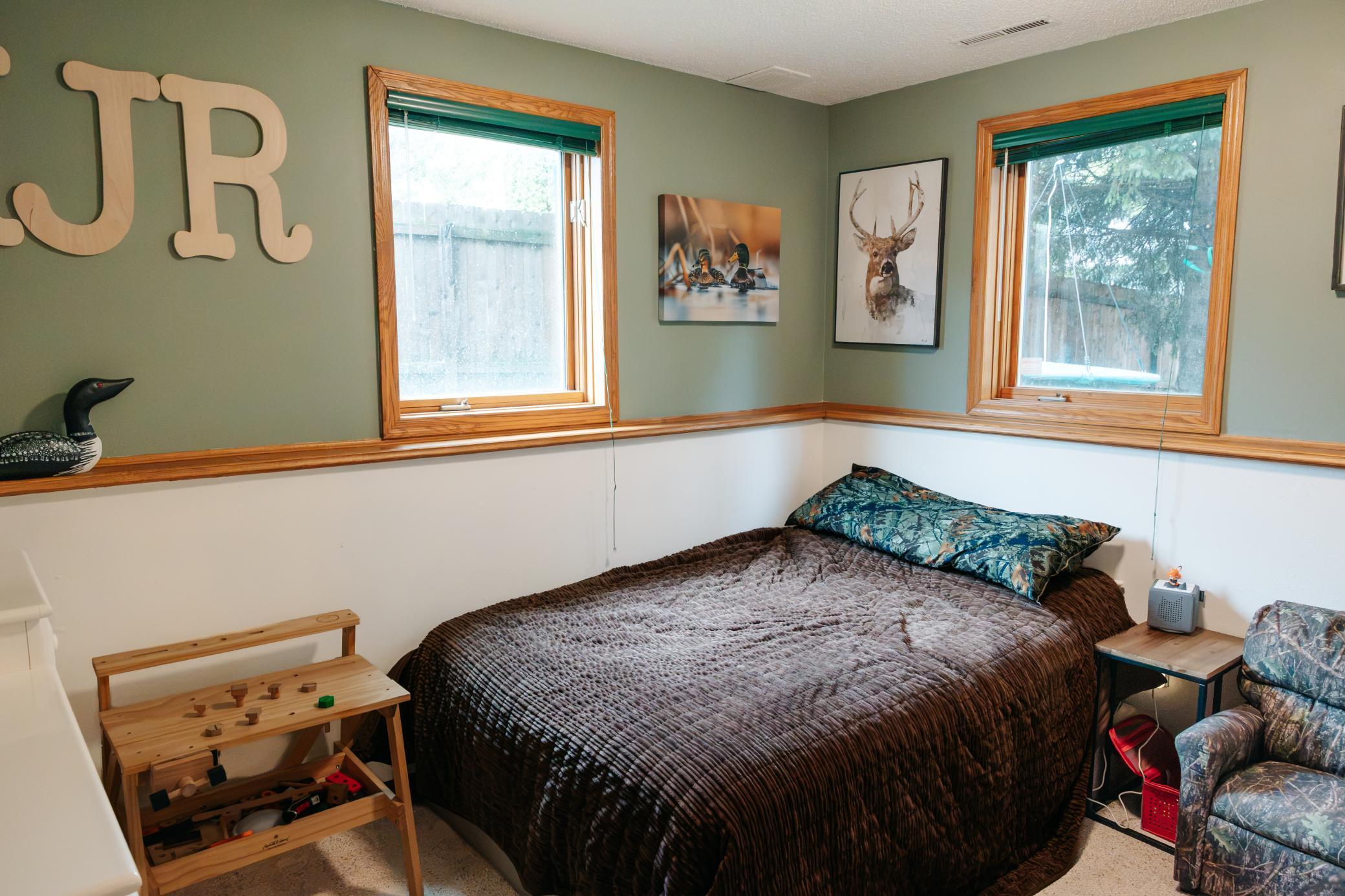 Furnished bedroom with a brown-quilted bed, wood-trimmed windows, and camouflage chair.