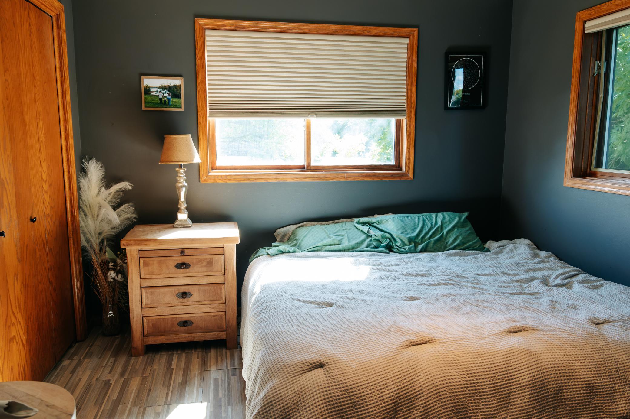 Furnished bedroom with a large bed, dark teal walls, and sunlit wood-trimmed windows.