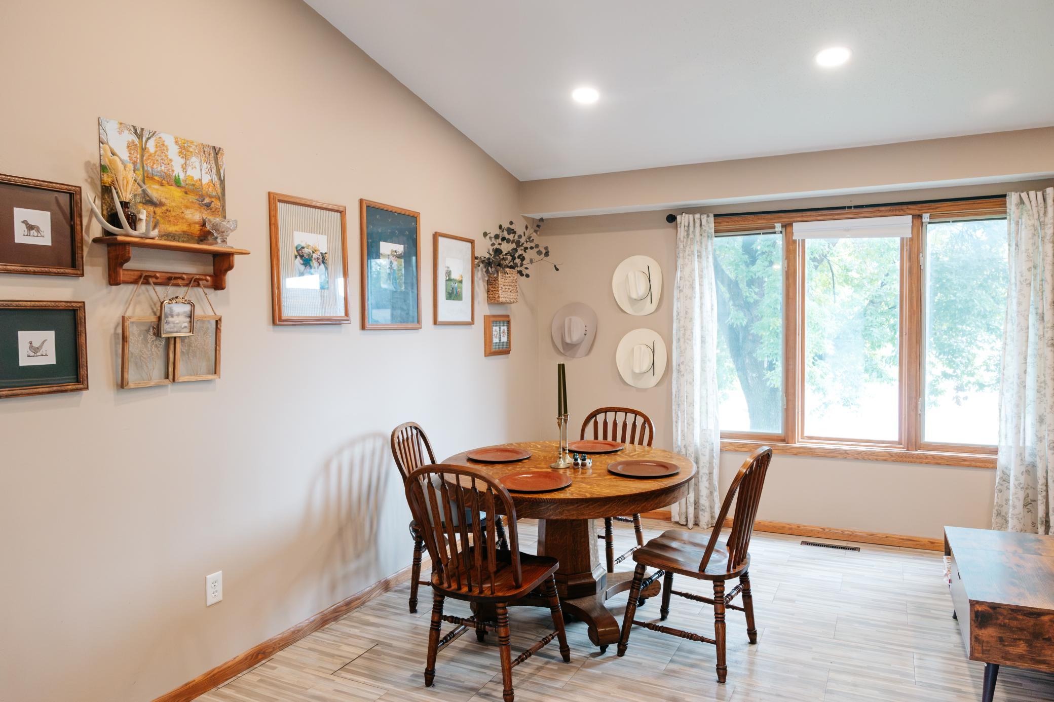 Dining room with a round wooden table, spindle-back chairs, gallery wall, and large curtained window.