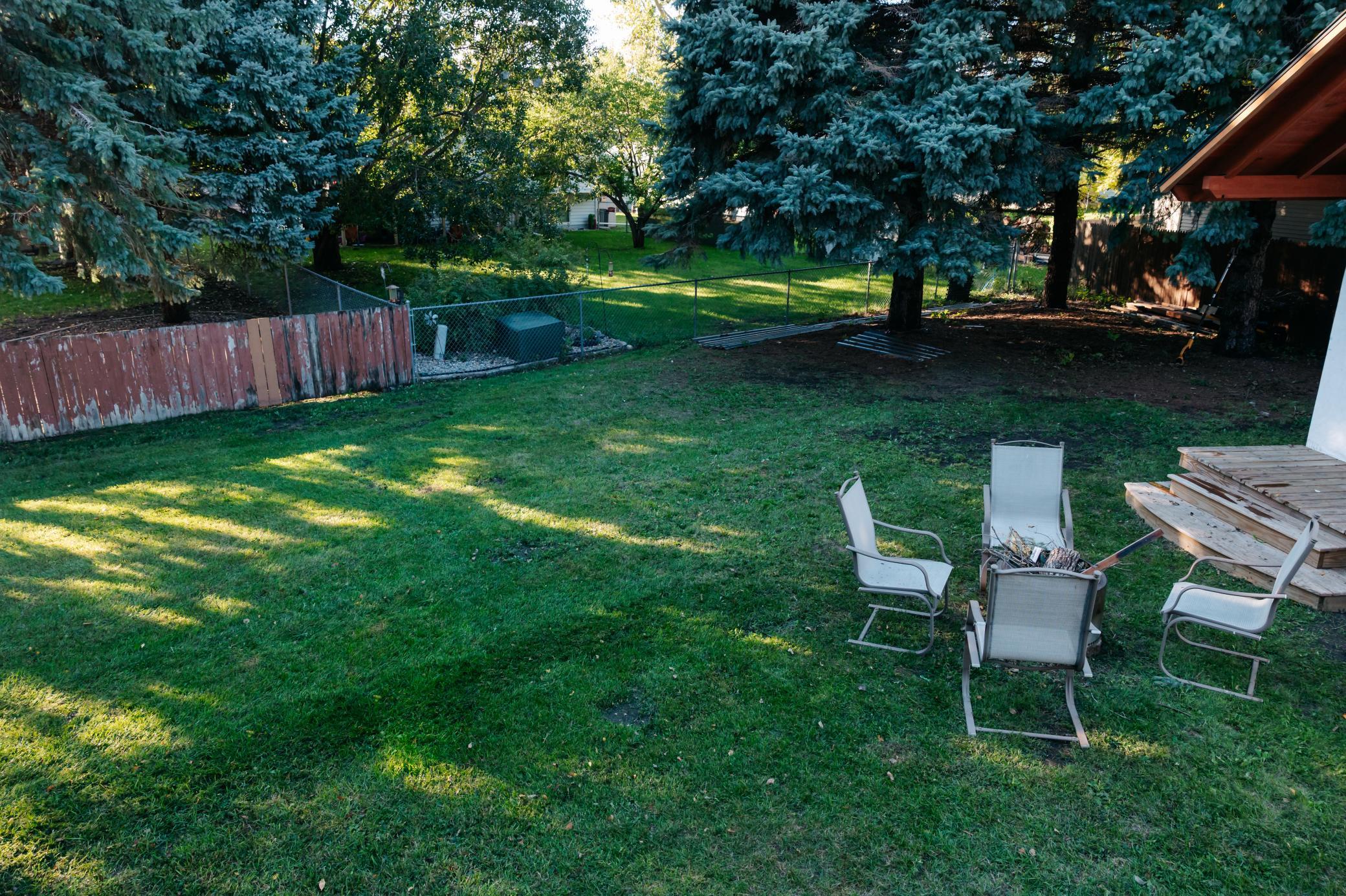 Backyard lawn beneath mature evergreens, with patio chairs around a fire pit and a raised wooden deck.