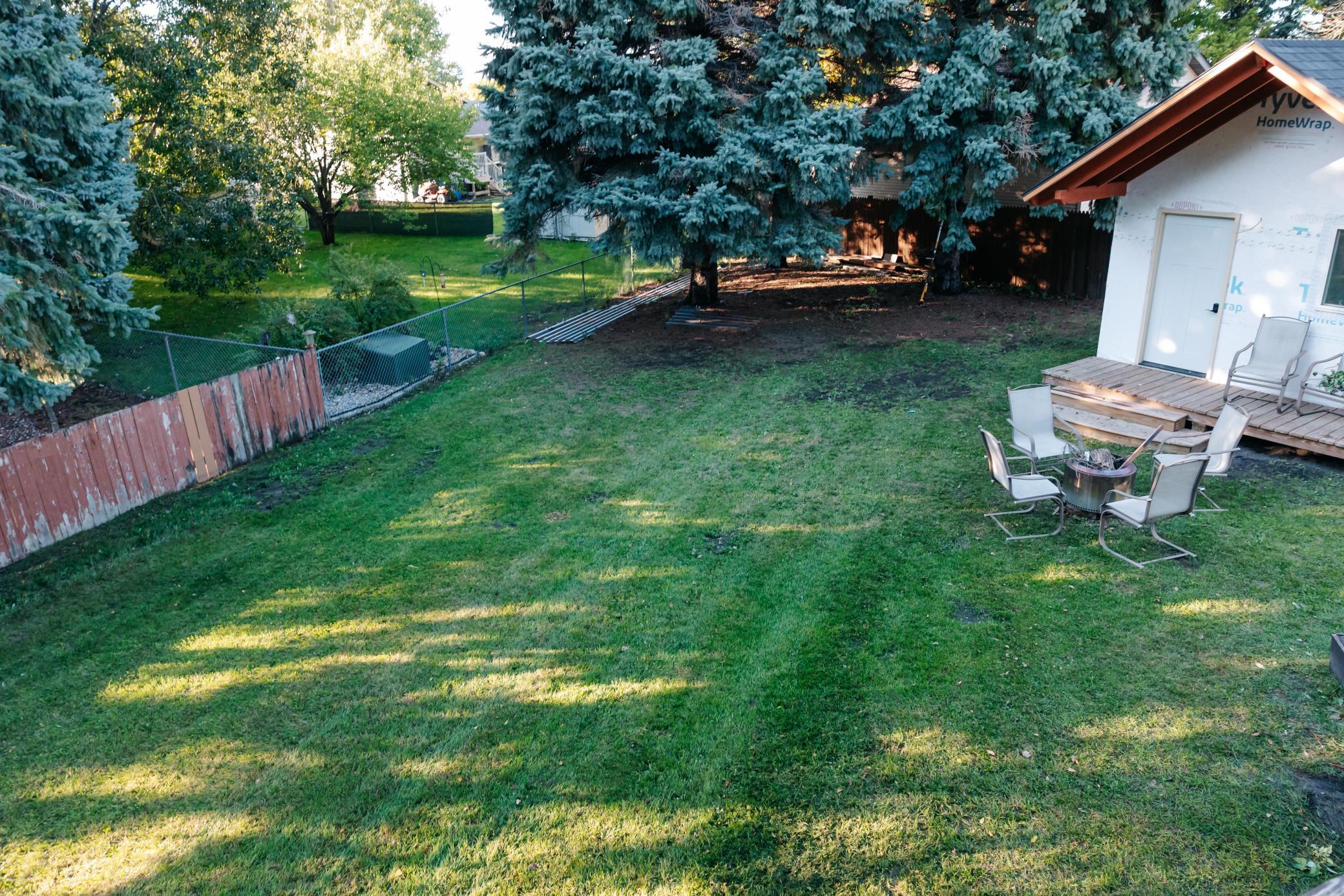 Shaded backyard with a lawn, evergreen trees, and patio seating around a fire pit.