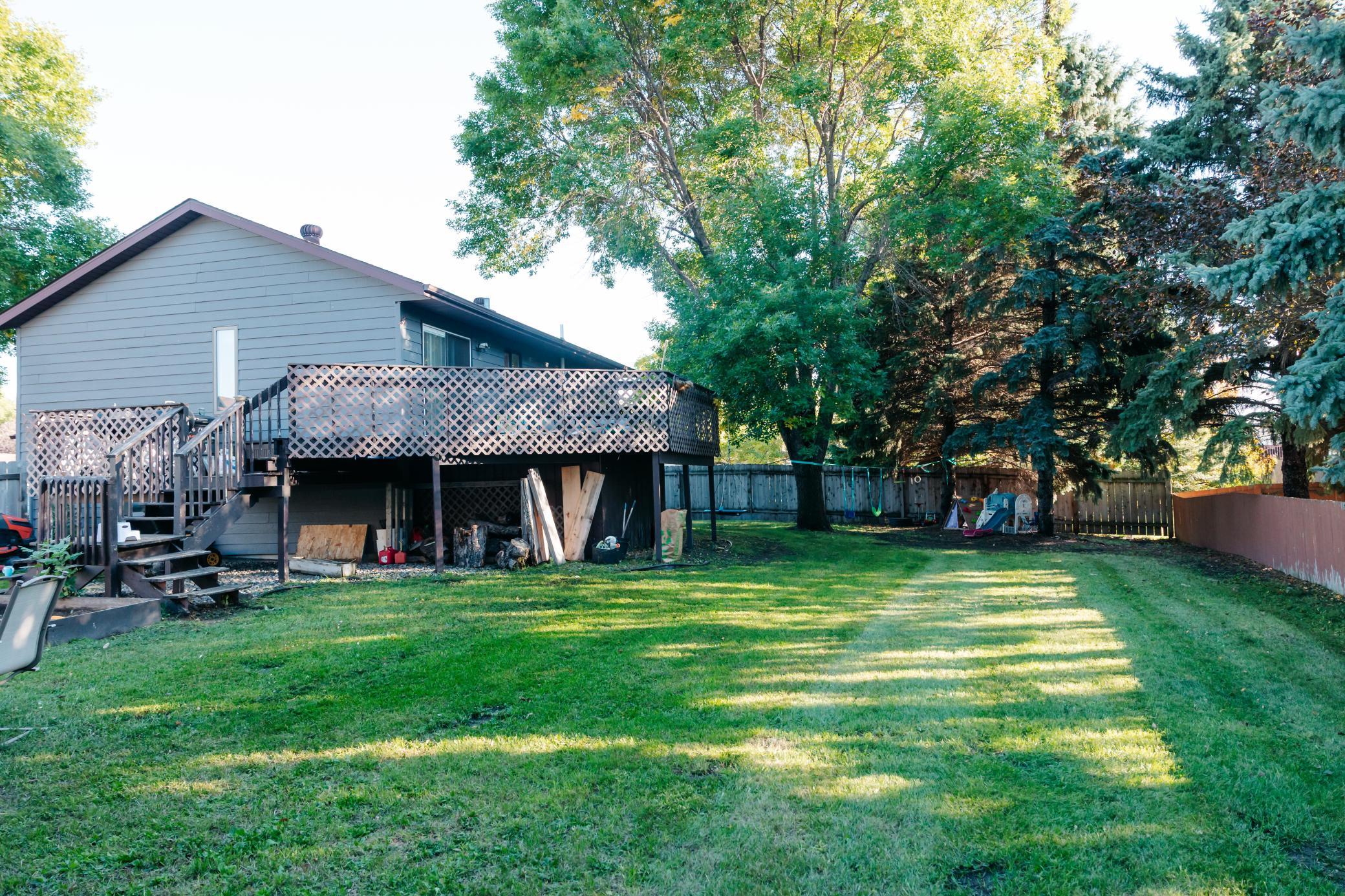Backyard with a raised deck, fenced lawn, mature trees, and children’s play equipment.
