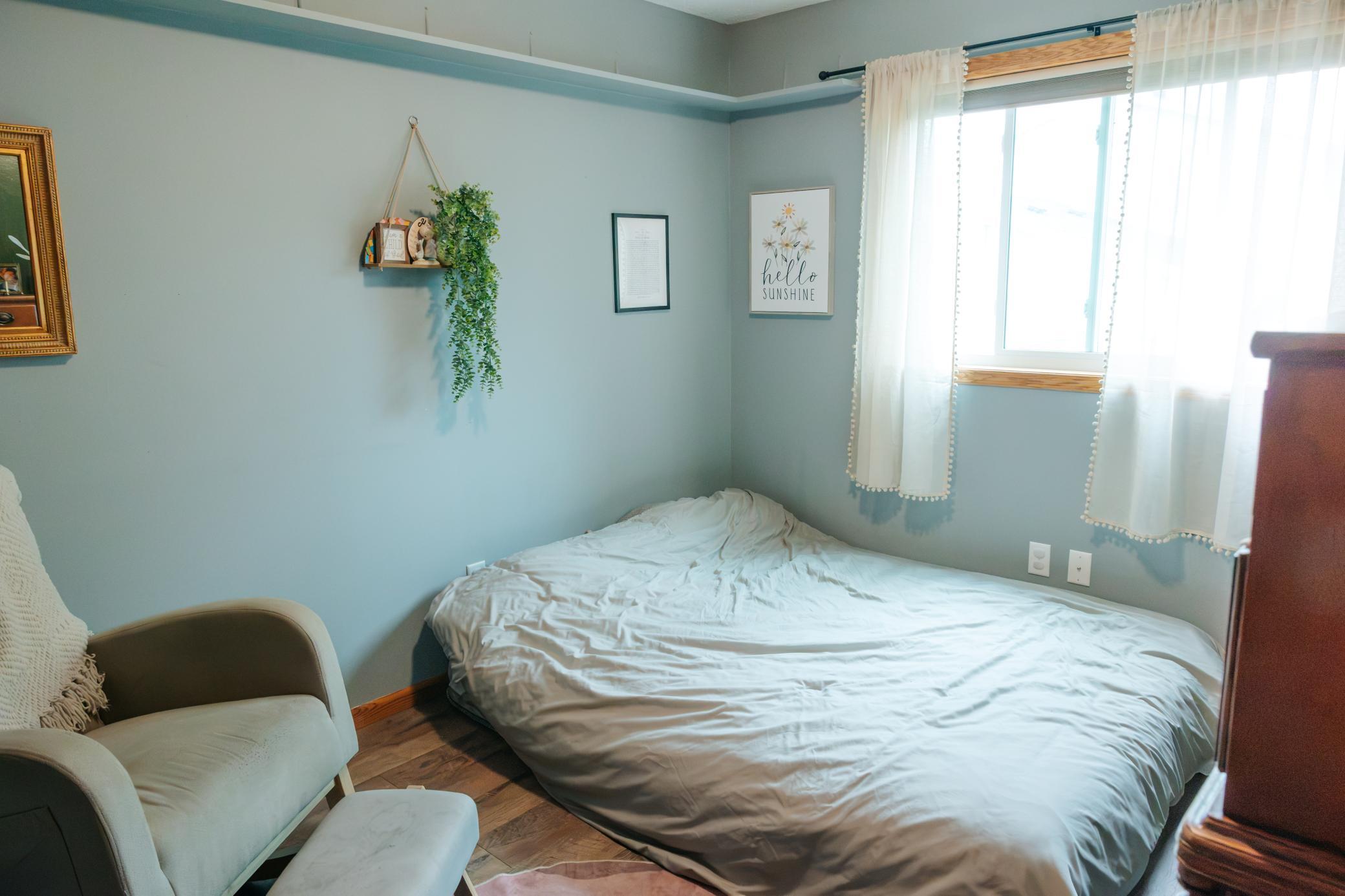 Bedroom with a low white bed, pale blue walls, sheer-curtained window, and upholstered chair.