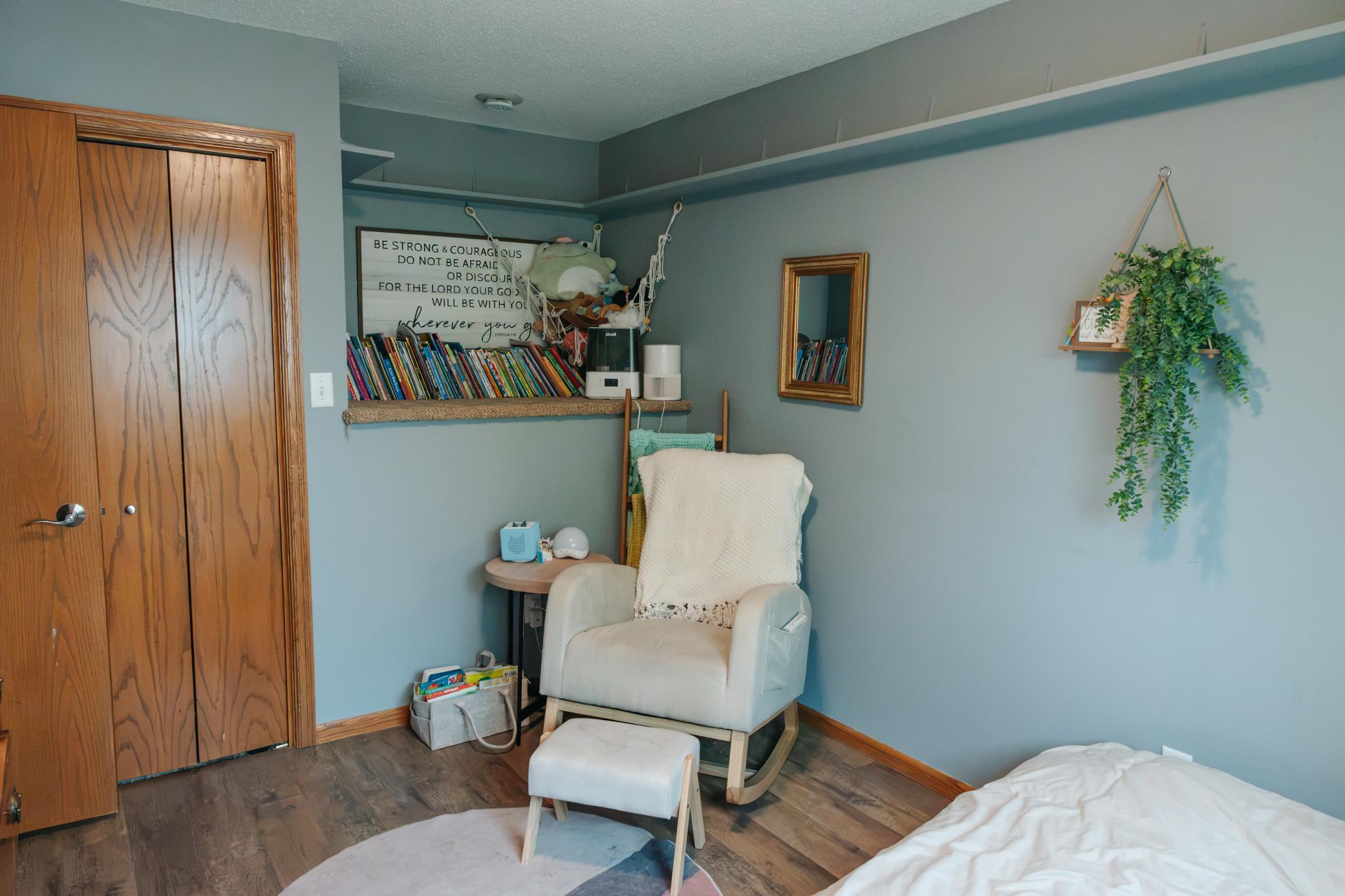 Bedroom with a cream rocking chair, footstool, and built-in shelf of children's books beside the bed.