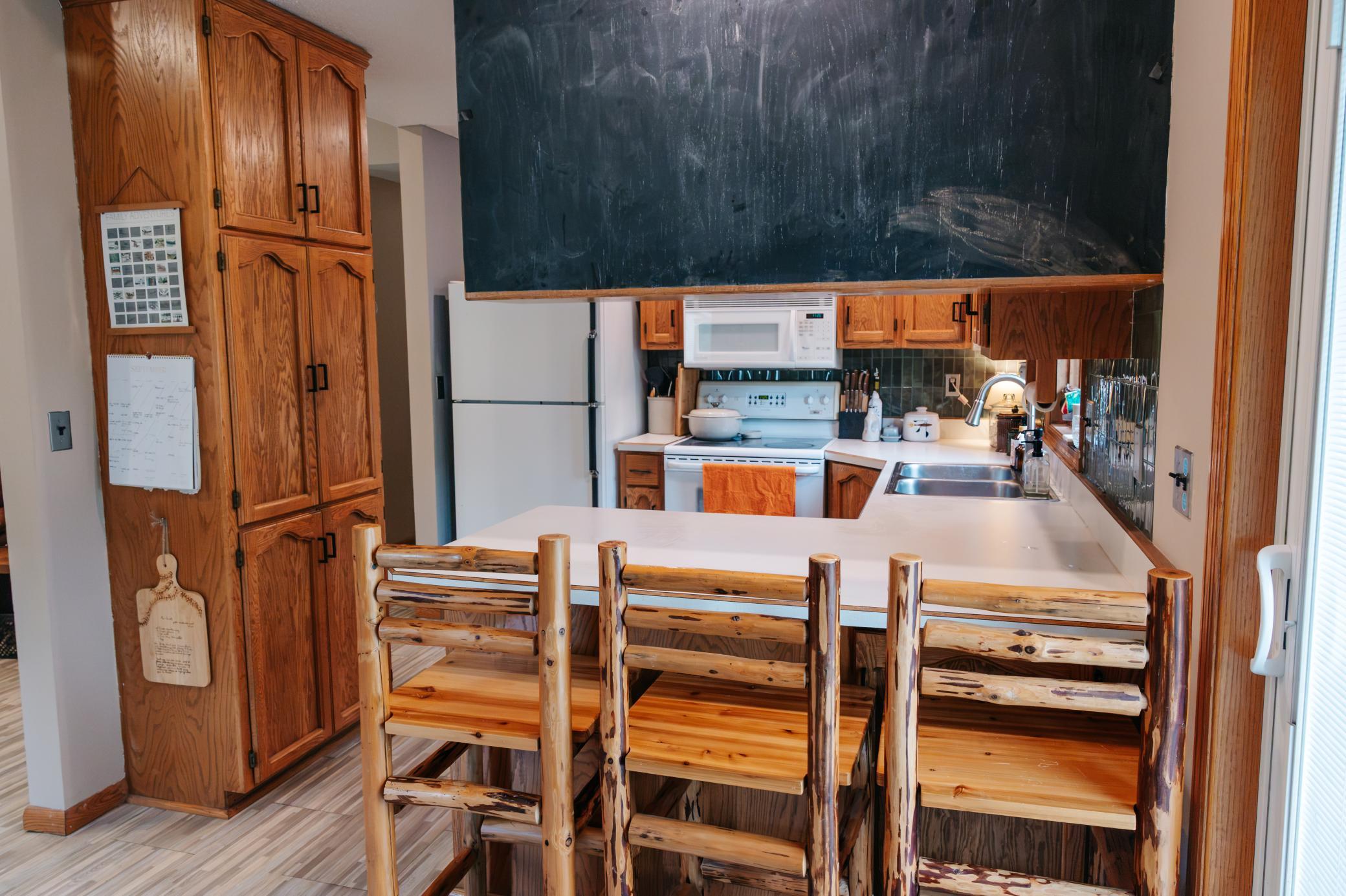 Kitchen with honey-colored wood cabinets, log-style bar stools, white counters, and a large chalkboard.