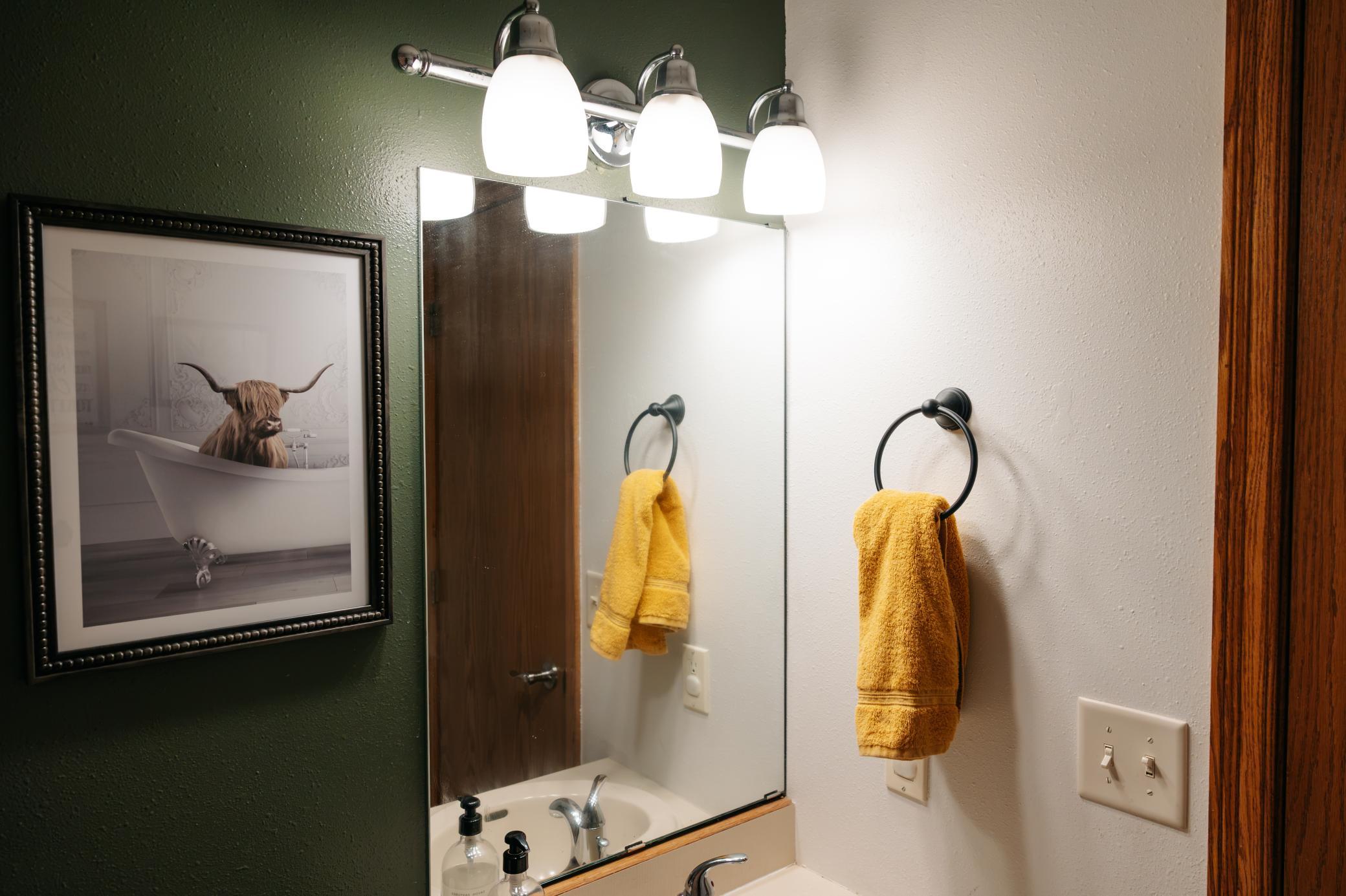 Bathroom vanity with a large mirror, three-light fixture, yellow towels, and framed Highland cow artwork.