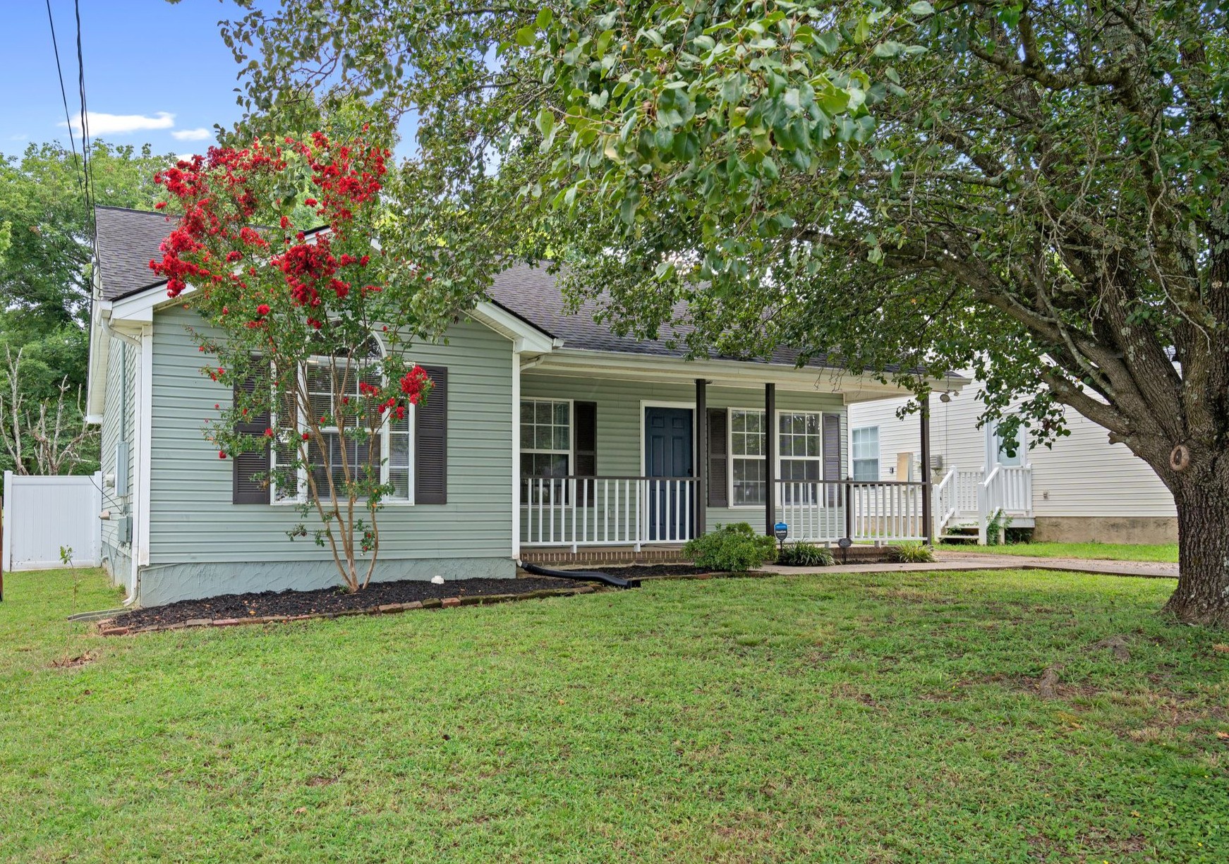 Sage-green single-story home with a covered porch, broad lawn, mature shade tree, and flowering crepe myrtle.