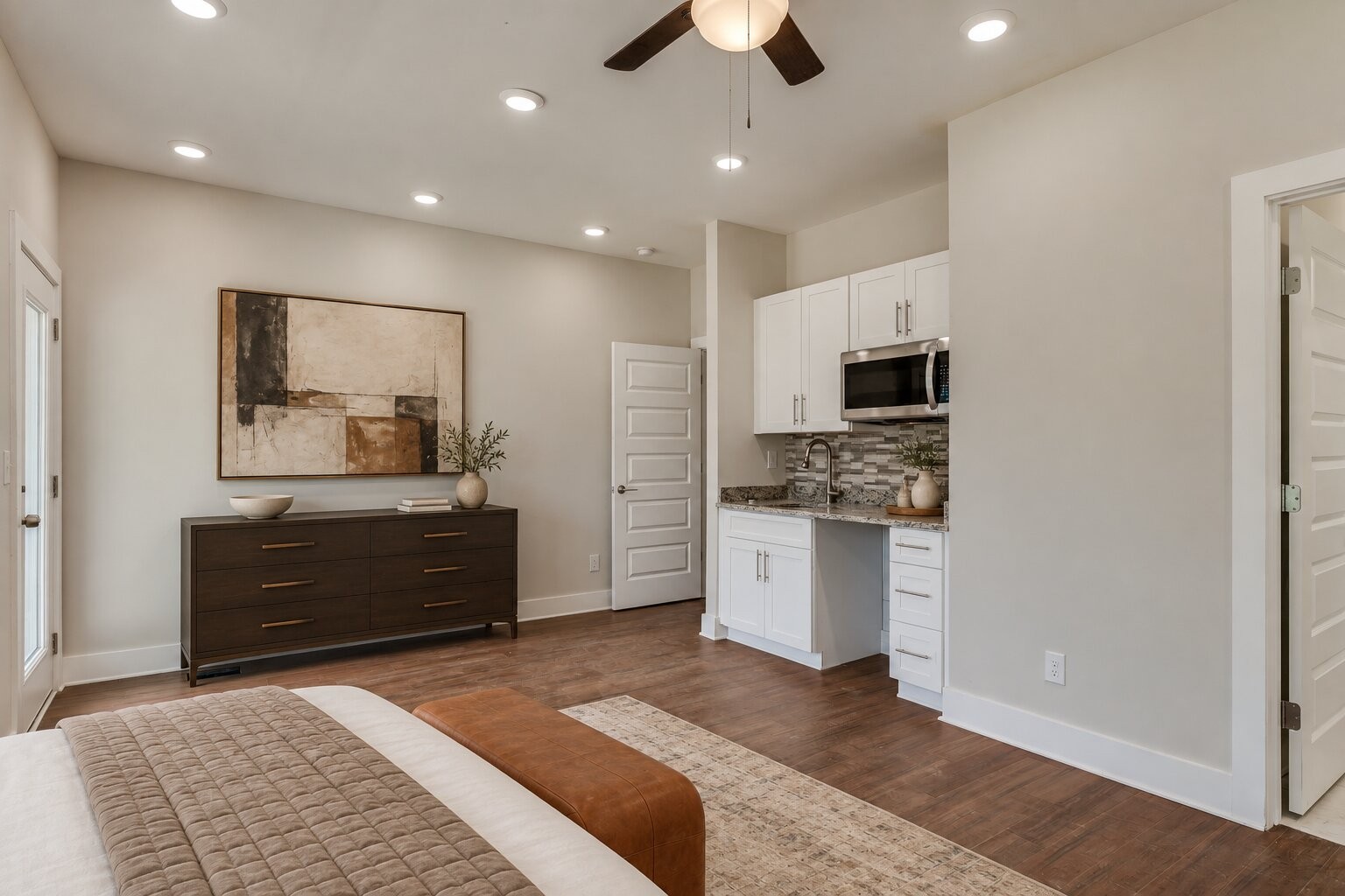 Bedroom with a compact white kitchenette, hardwood floors, and a dark dresser beneath abstract artwork.