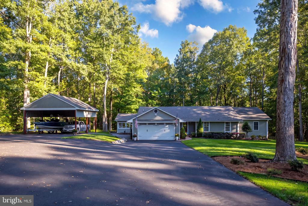 Gray single-story home with a garage, wooded yard, and boat under a covered carport.