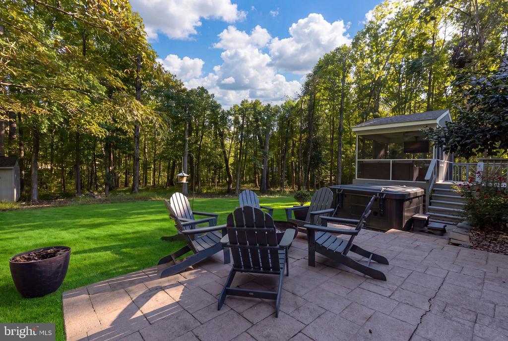 Stone patio with Adirondack chairs and hot tub beside a lawn and wooded yard.