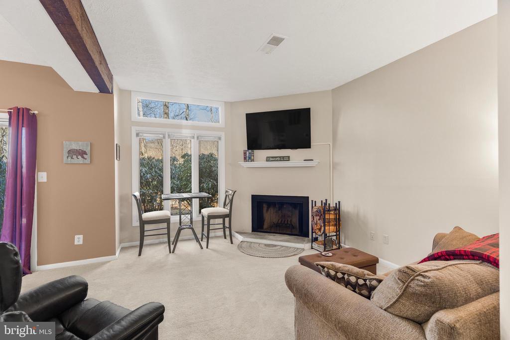 Furnished living room with a fireplace, exposed ceiling beam, and glass doors overlooking shrubs.