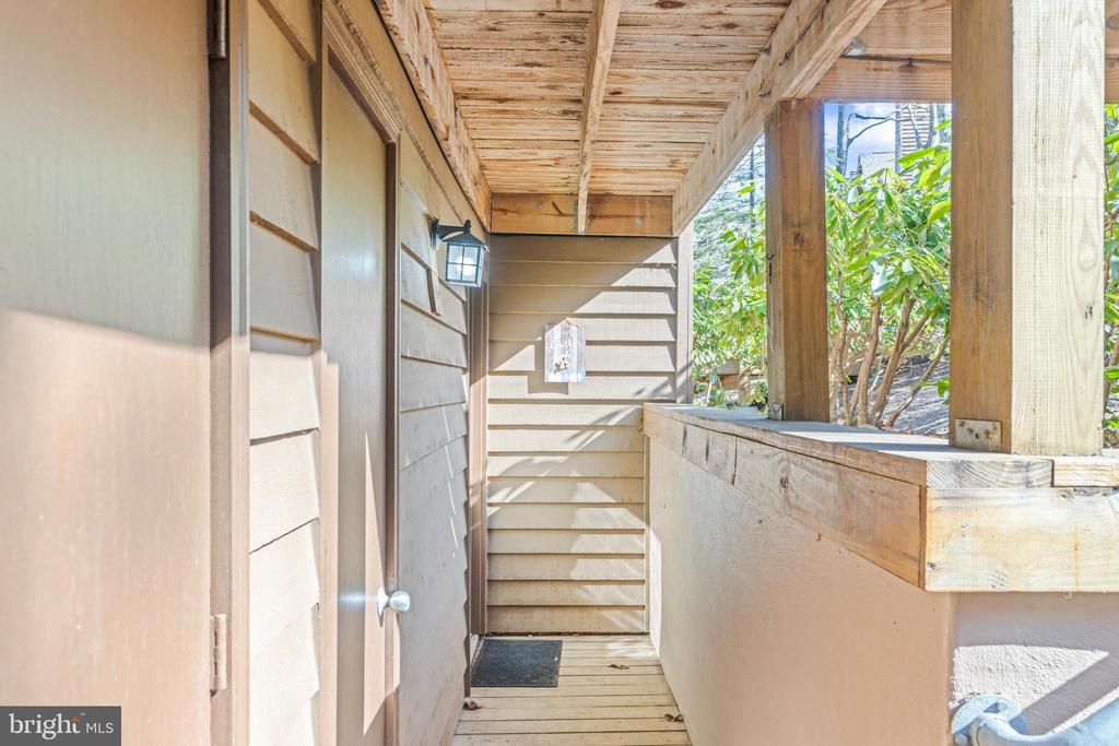 Covered porch with a wooden ceiling, tan siding, and a wall lantern beside the entry door.
