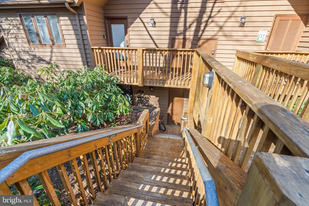 Wooden stairs lead to an elevated deck beside a tan-sided home, with shrubs along the railing.